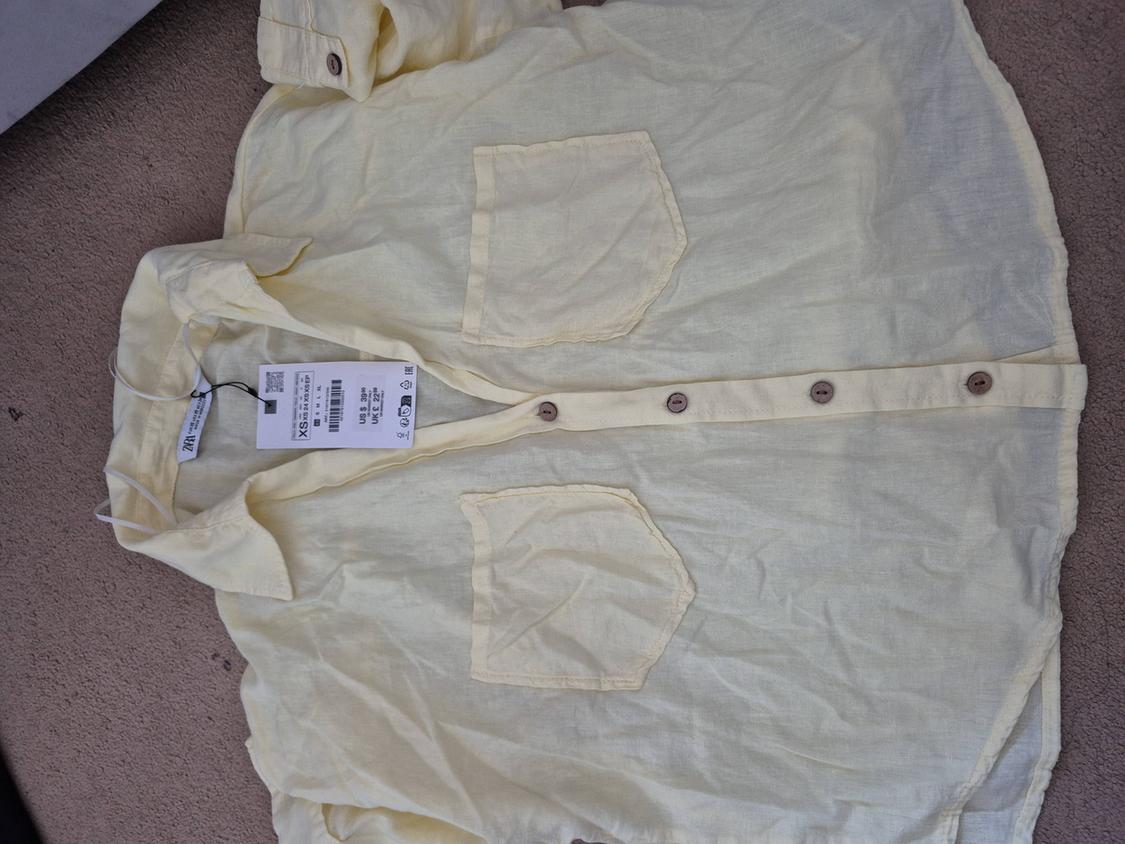 Yellow linen shirt with two front pockets and brown buttons, size XS label visible, on a beige surface.