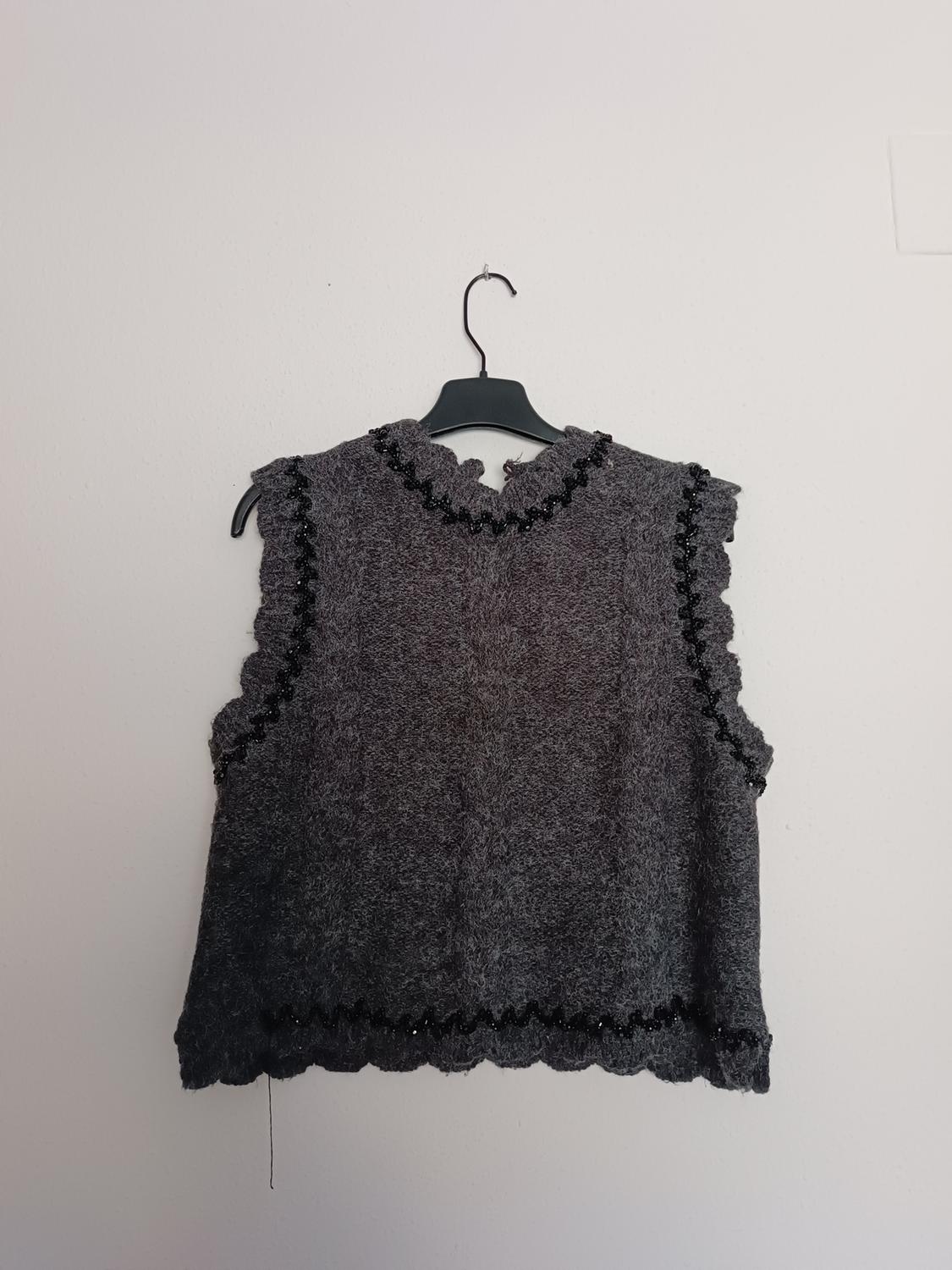 Sleeveless grey waistcoat with wavy edges and black lace details, hanging on a black hanger against a white background.
