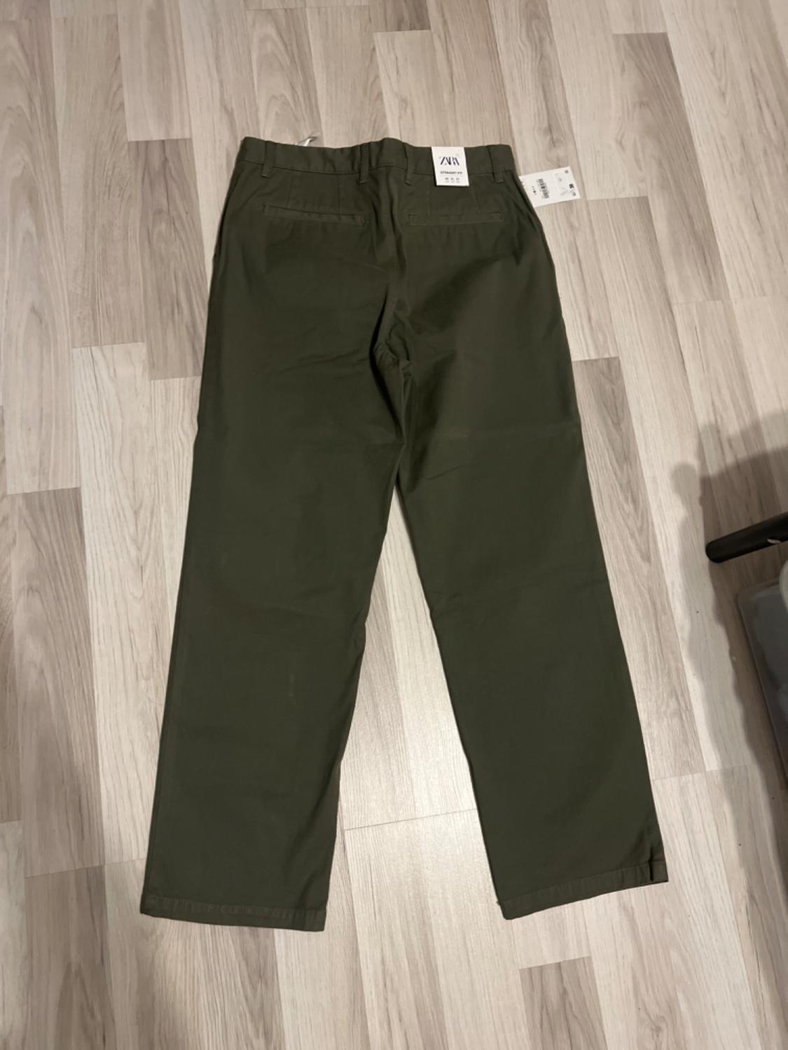 Straight-cut green trousers on a light wooden floor. Labels visible on the back of the trousers.
