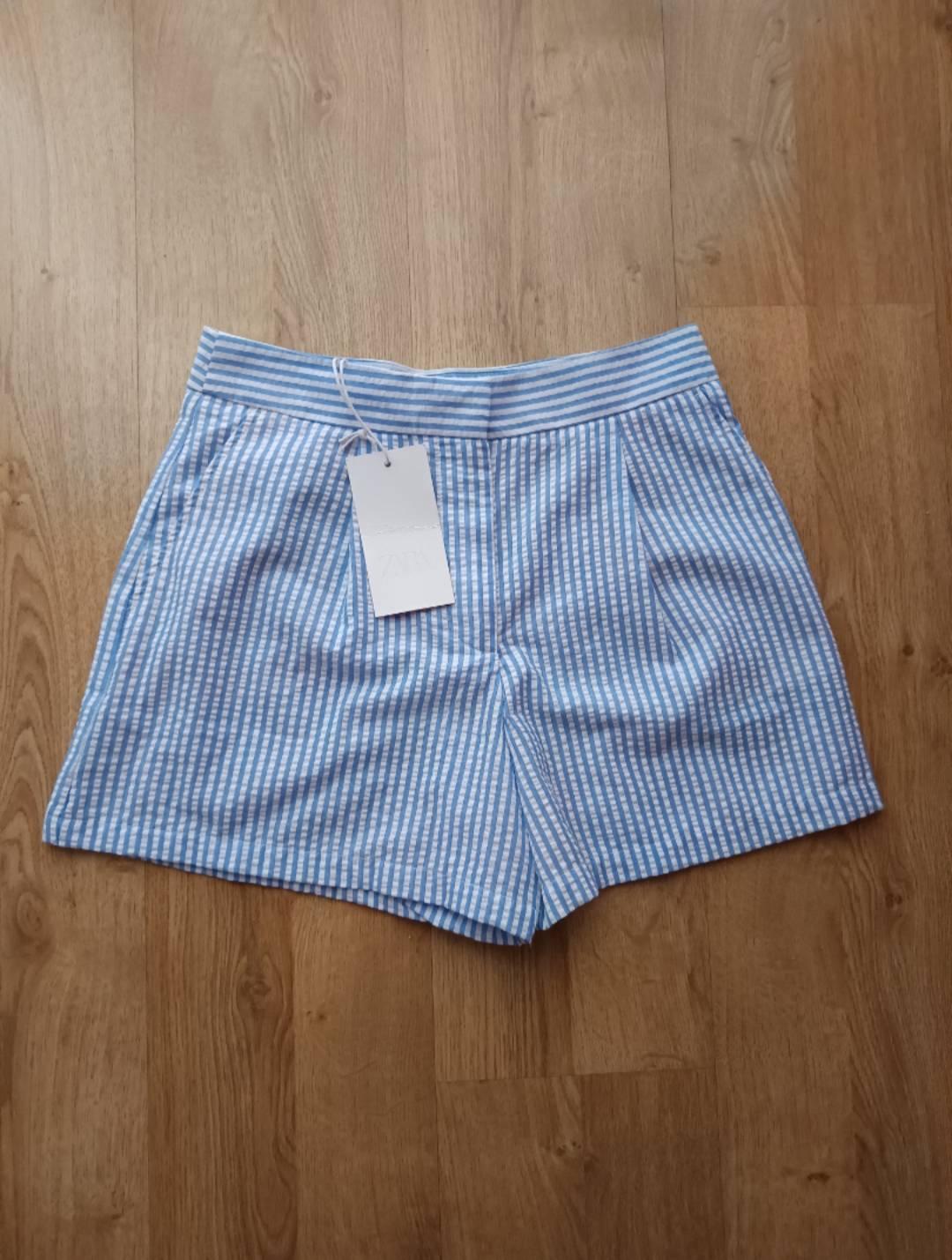 Blue and white striped tailored shorts with ZARA label, on a wooden floor. Casual flat lay composition.