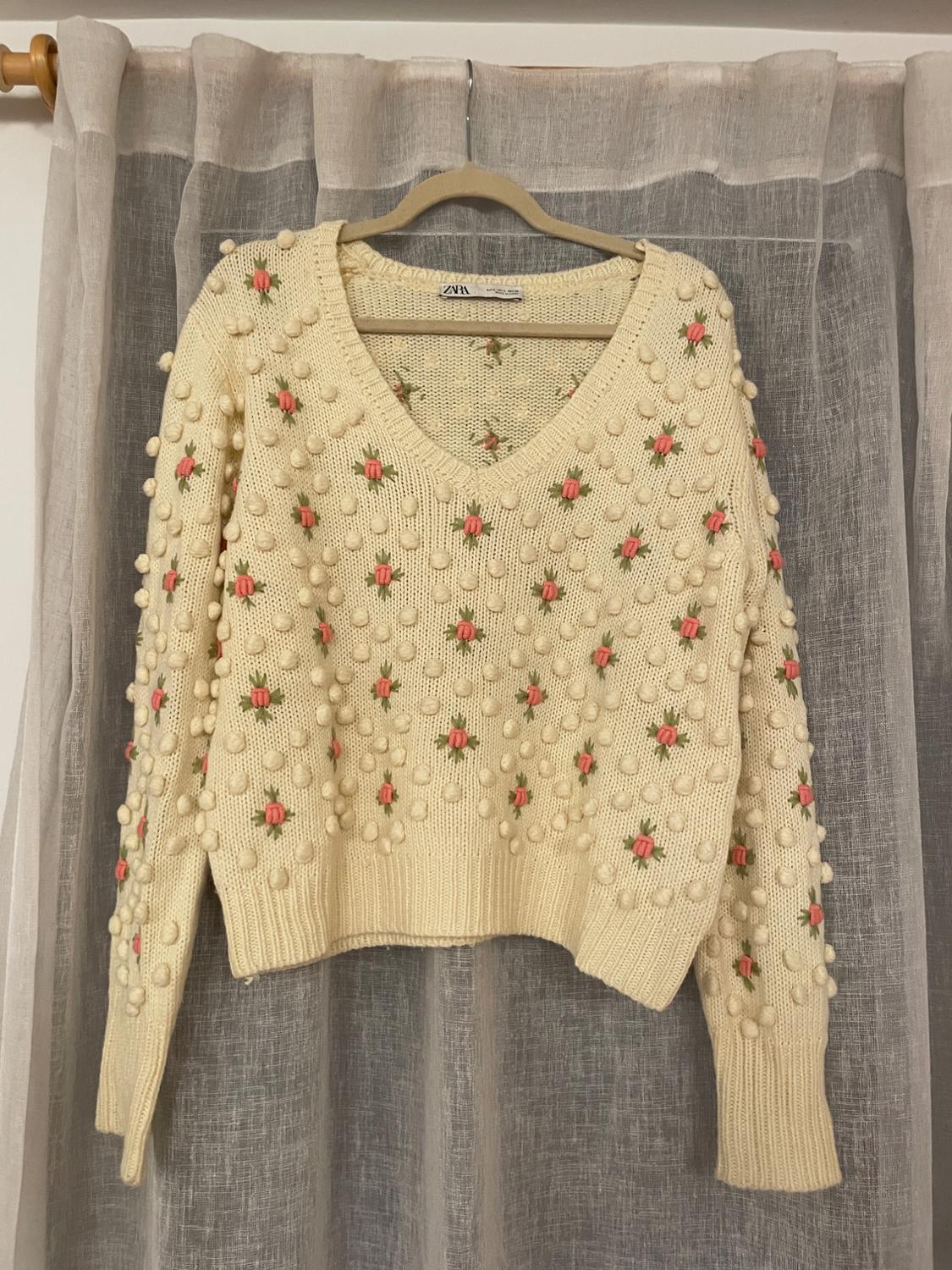Cream sweater with pompoms and pink floral embroidery, hanging on a hanger in front of a translucent gray curtain.