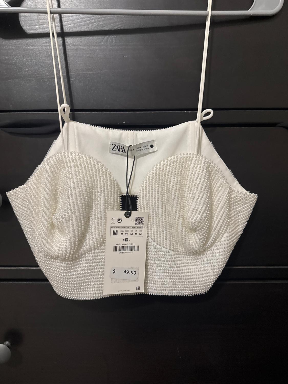 White camisole with thin straps and pearl texture hanging on a hanger. Visible tag with price of $49.90 and size M.