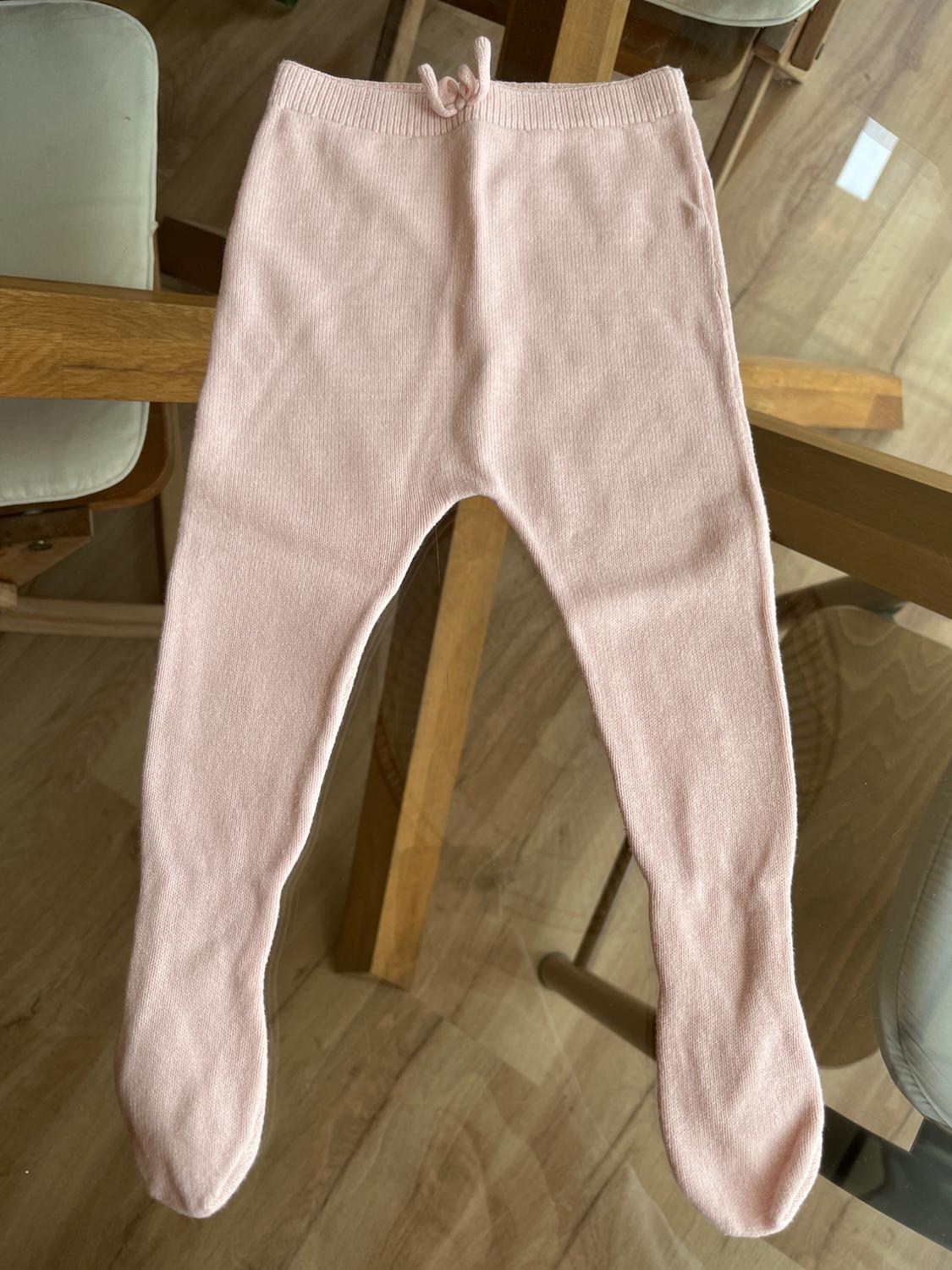 Light pink baby trousers with feet and a tie at the waist, on a glass table with a light wooden background.