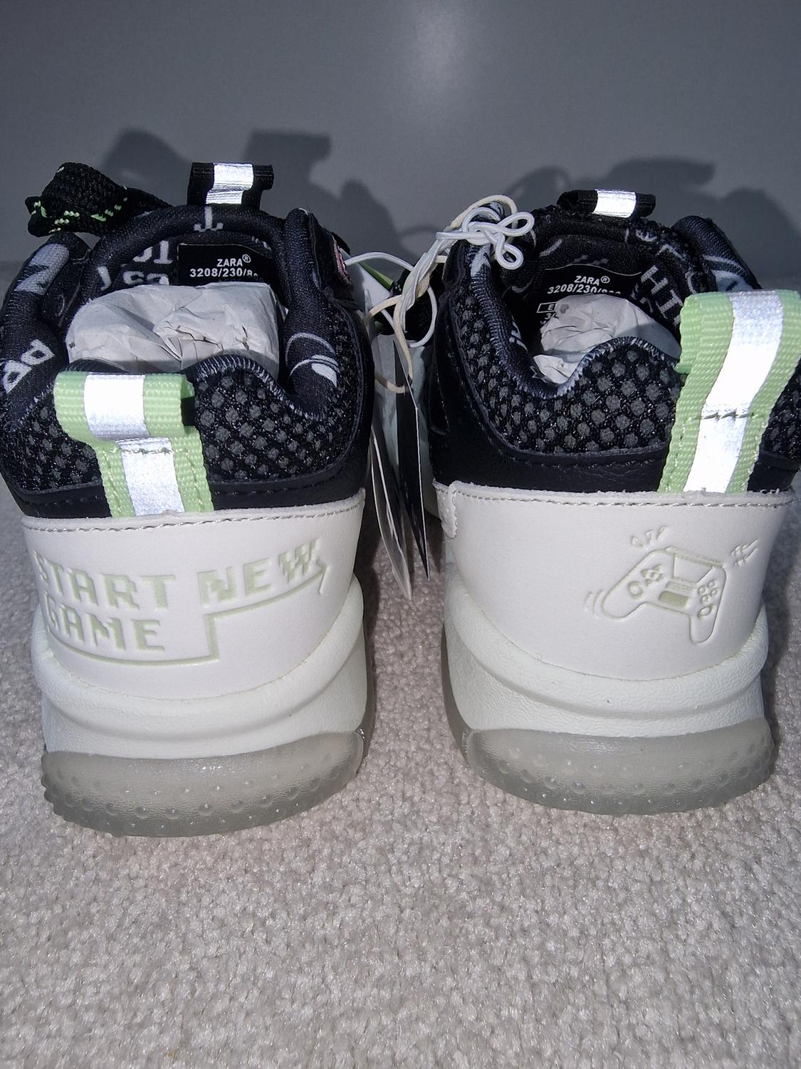Black and white trainers with green detailing, rear view. Text "START NEW GAME" and controller design on the heel.