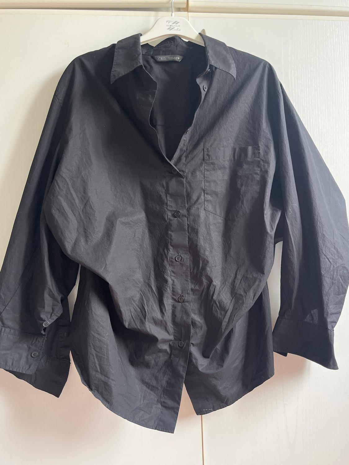 Black long-sleeved shirt hanging on a white hanger, with a light wall background.