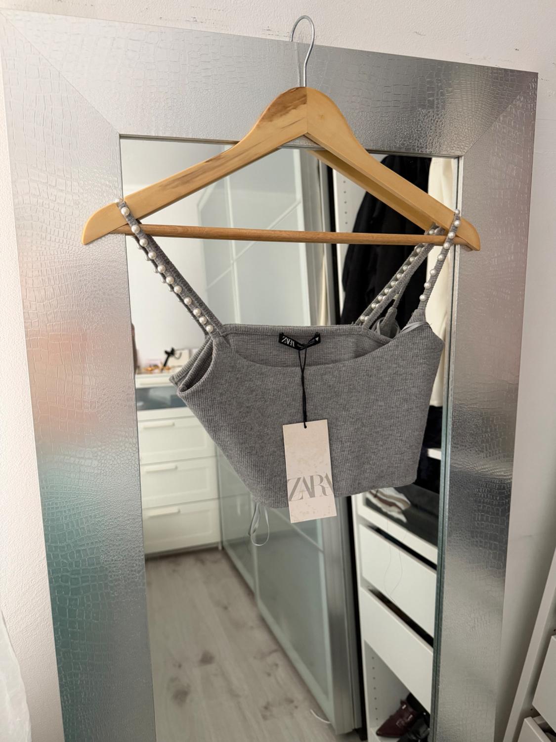 Grey top with pearl straps hanging on a wooden hanger in front of a mirror. Visible label with text "ZARA".