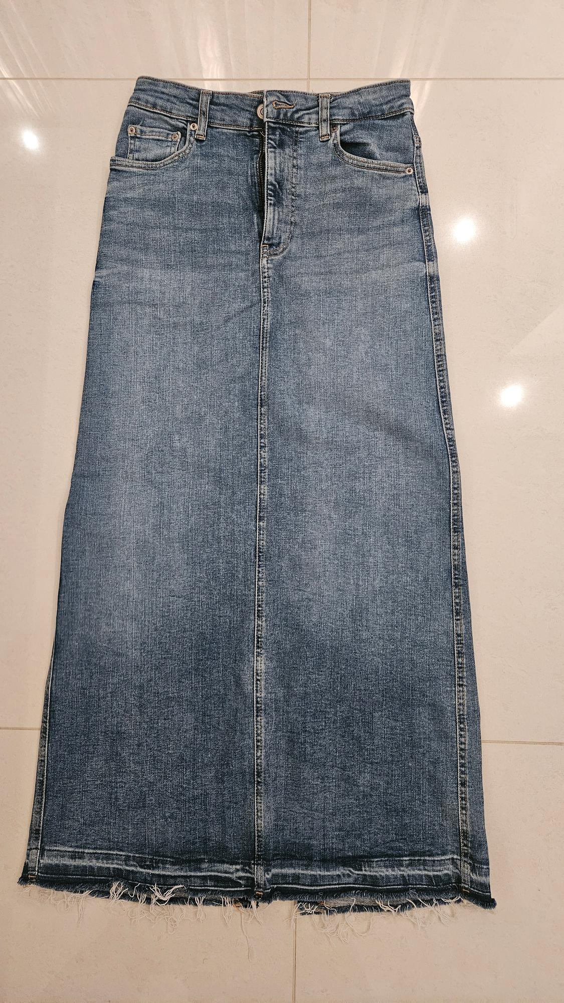 Blue denim maxi skirt with a frayed hem, placed on a light-coloured floor.