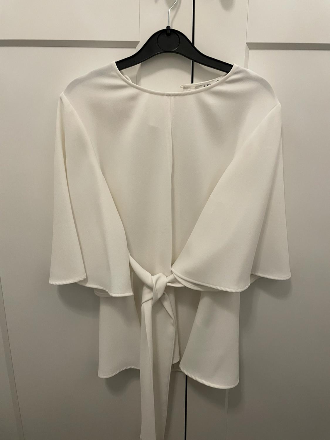 White short, wide-sleeved blouse with a round neck and front knot, hanging on a black hanger in front of a white wardrobe.