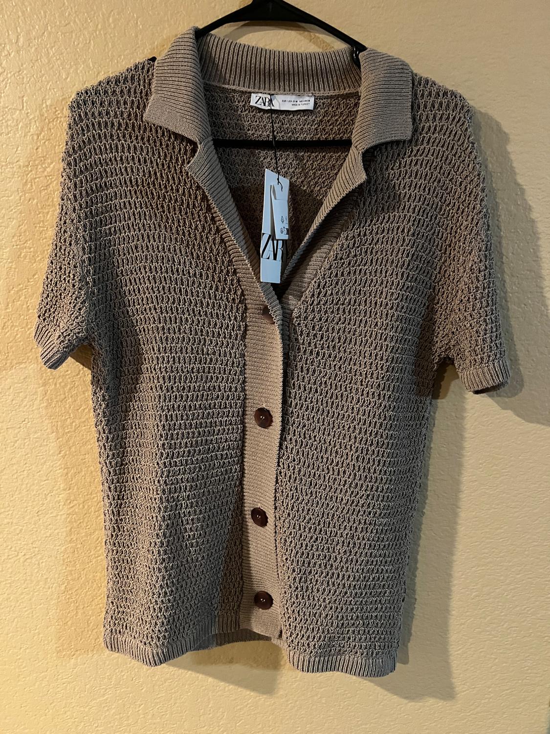 Brown knitted cardigan with dark buttons and folded collar, hanging on a hanger against a beige wall. Label visible.