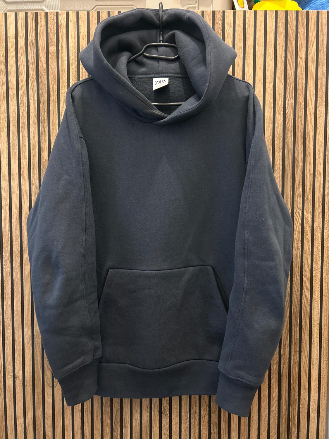 Blue hoodie hanging on a black hanger, against a background of wooden panels with vertical lines. Label visible.