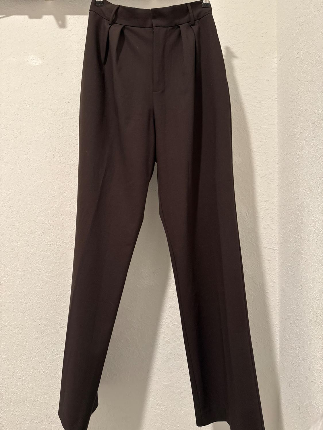 Black trousers hanging on a hanger against a white background. Pleated waist design and straight cut.