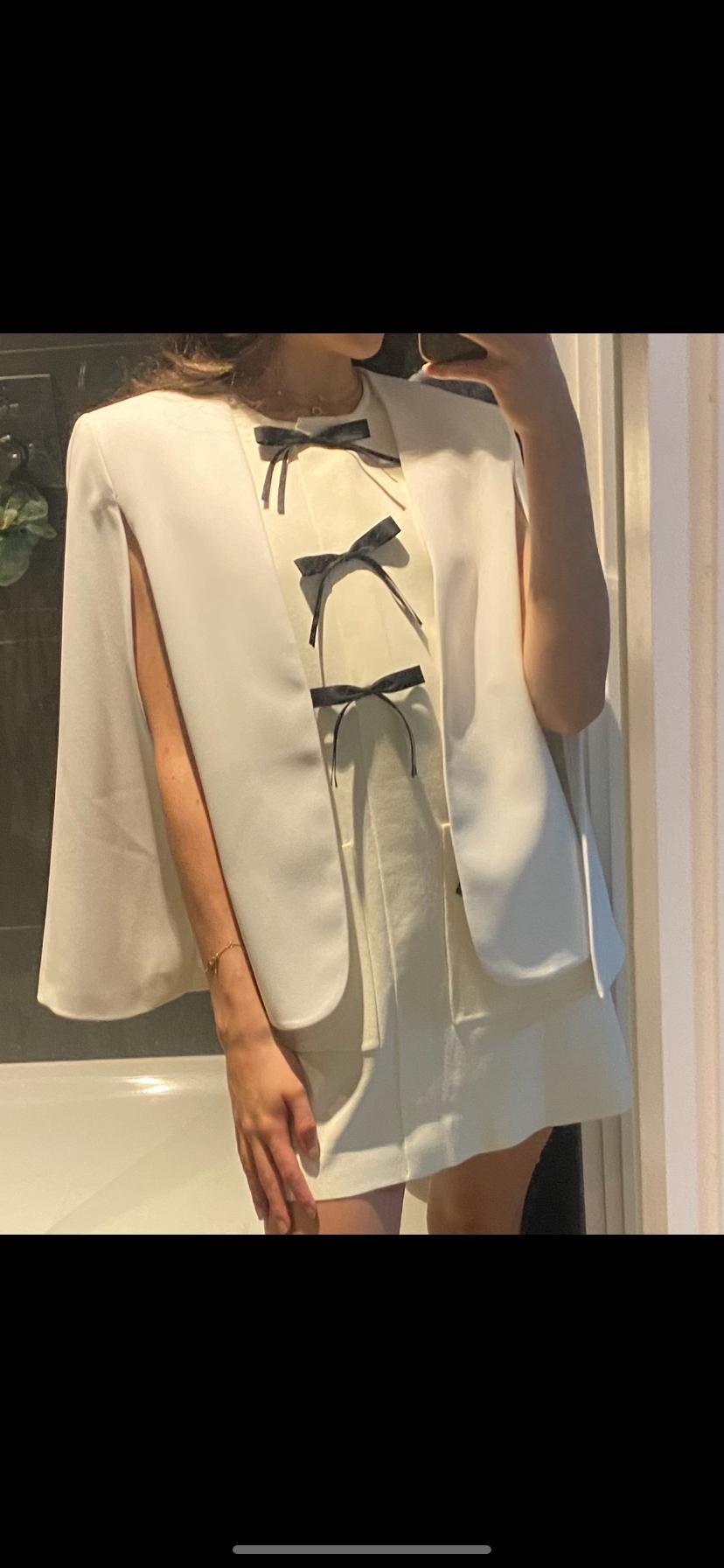 Mirror selfie of a woman wearing a short white dress decorated with three black bows and a white cape-style blazer.