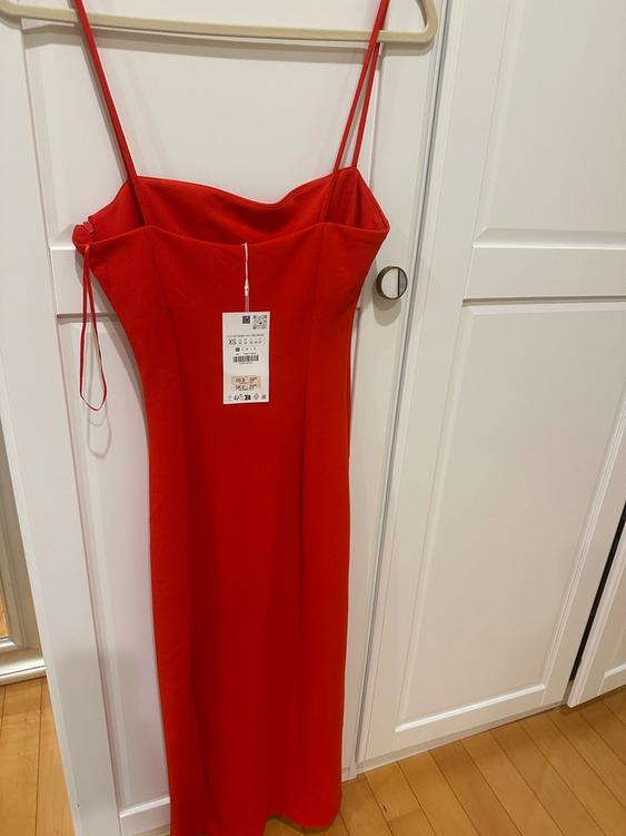 Red spaghetti strap dress hanging on a hanger, with a visible tag. White door background and light wood floor.