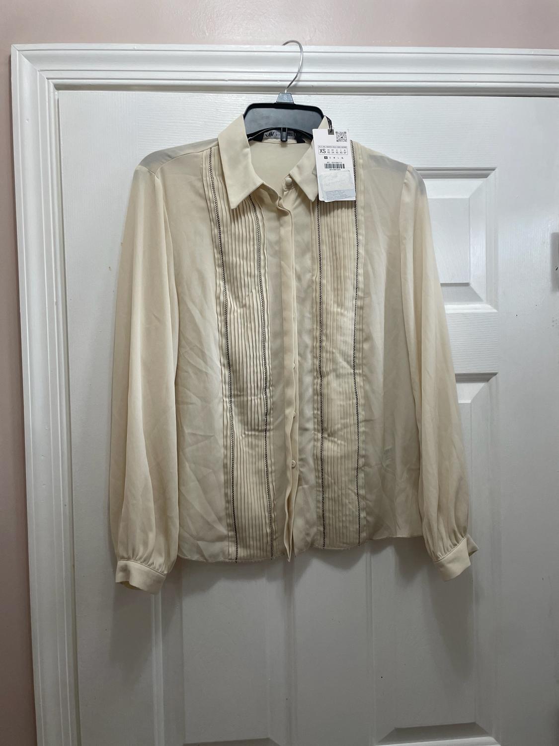 Cream-colored long-sleeve blouse with vertical pleats and dark seams on the front, hanging on a hanger over a white door.