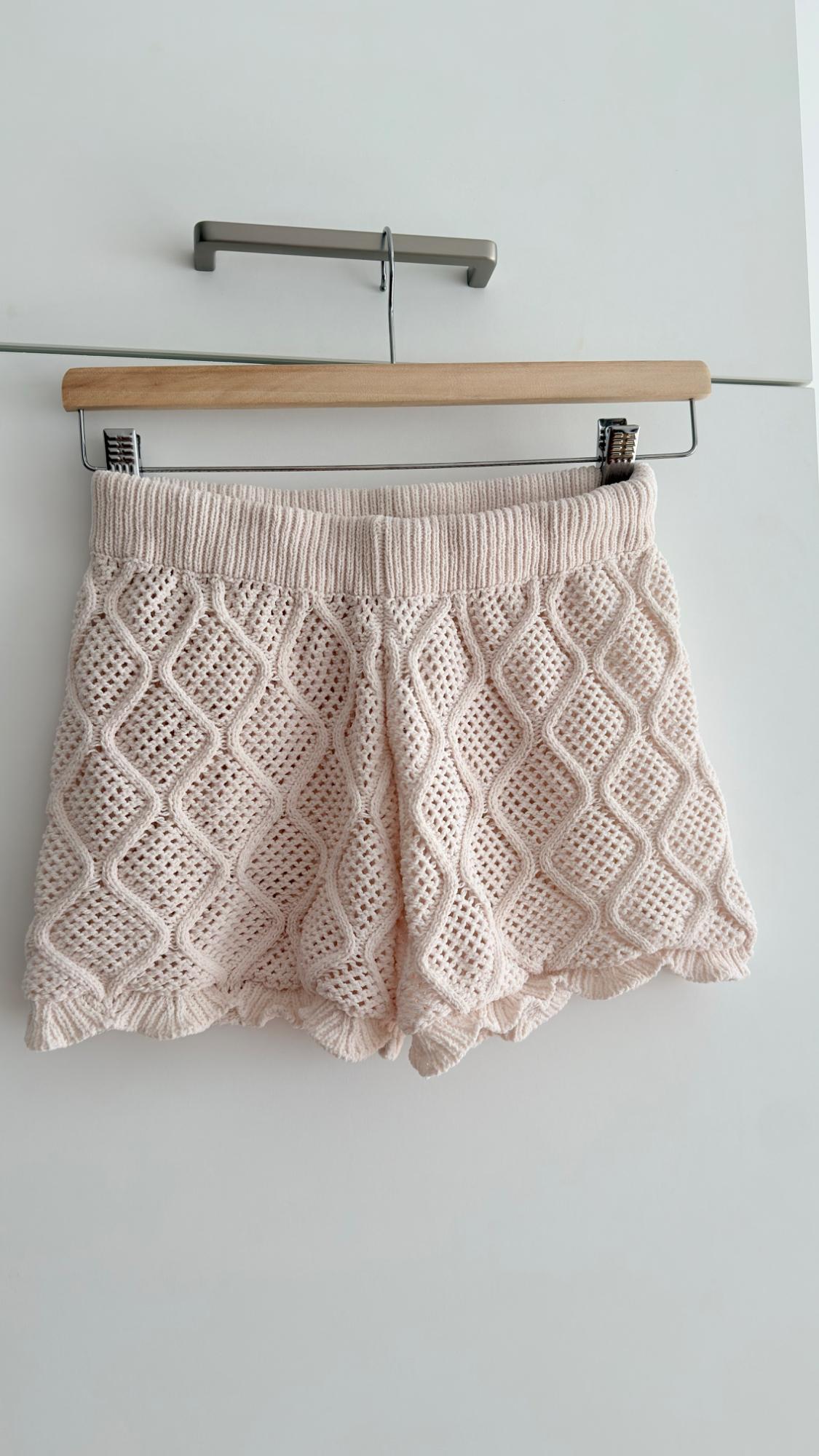 Beige crochet shorts hanging on a wooden hanger against a white background. Diamond design and elasticated waist.