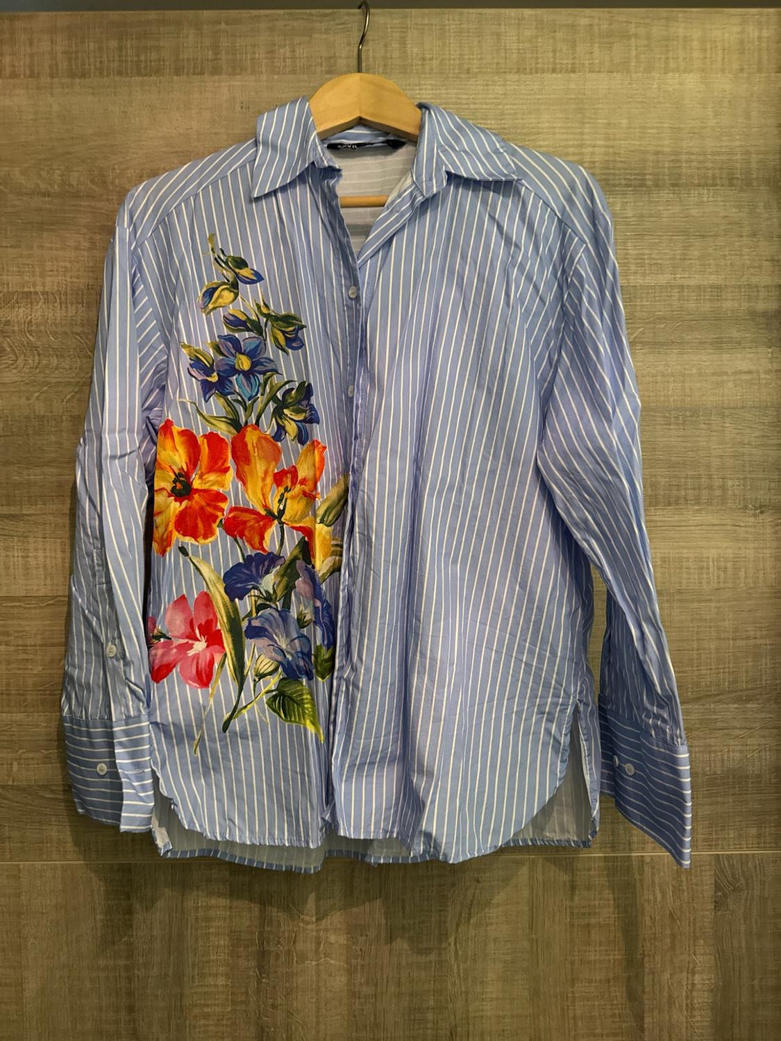 Blue and white striped long-sleeved shirt with a colourful floral print, hanging on a hanger against a wooden background.