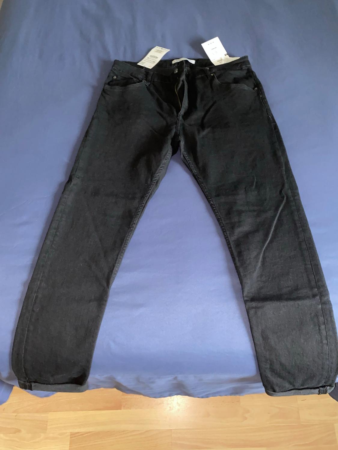 Black denim pants with tags, laid out on a blue sheet. The zipper is slightly open, and the cuffs are folded up.