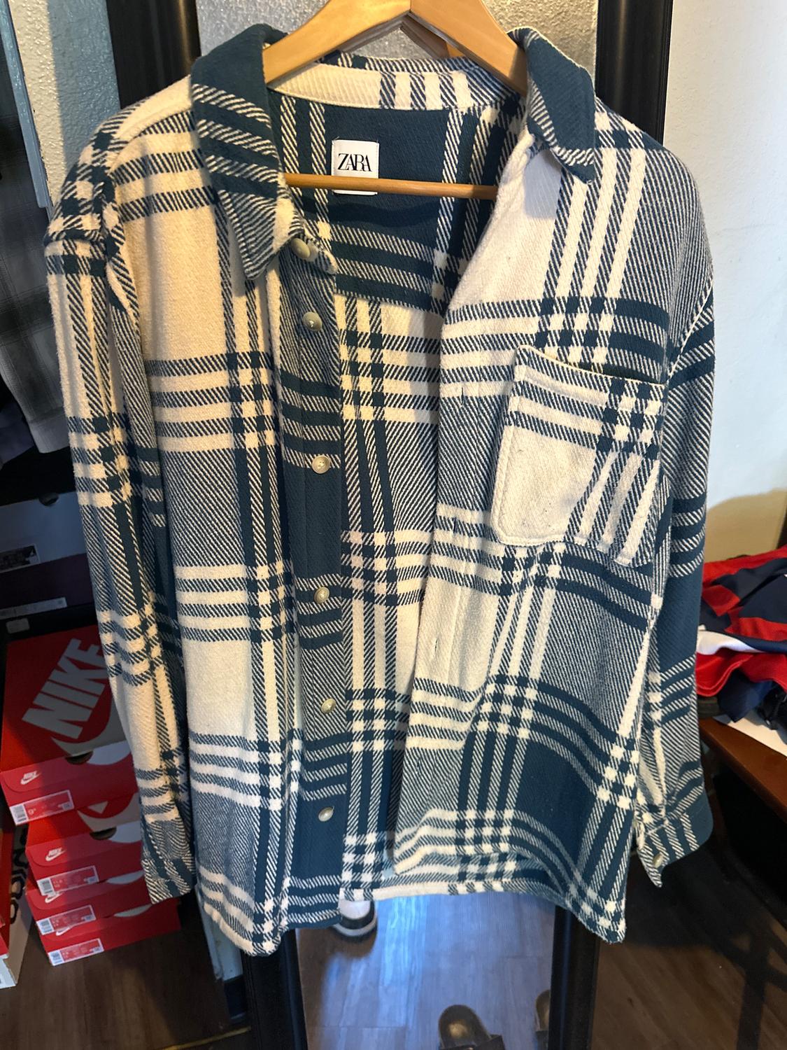 Blue and white checked shirt hanging on a wooden hanger, with a visible Zara label. Background with shoe boxes.