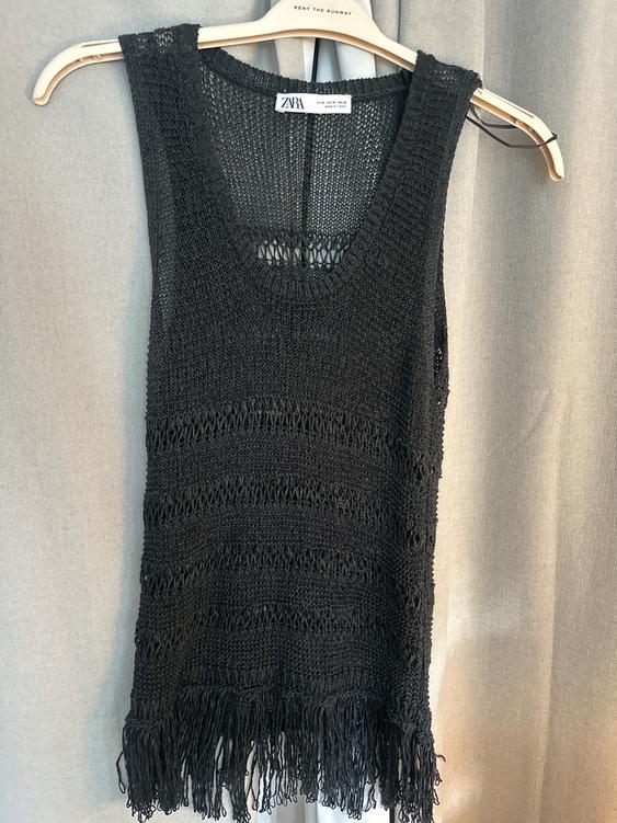 Black knit top with fringe at the hem, hanging on a wooden hanger. Visible tag: "ZARA". Gray curtain background.
