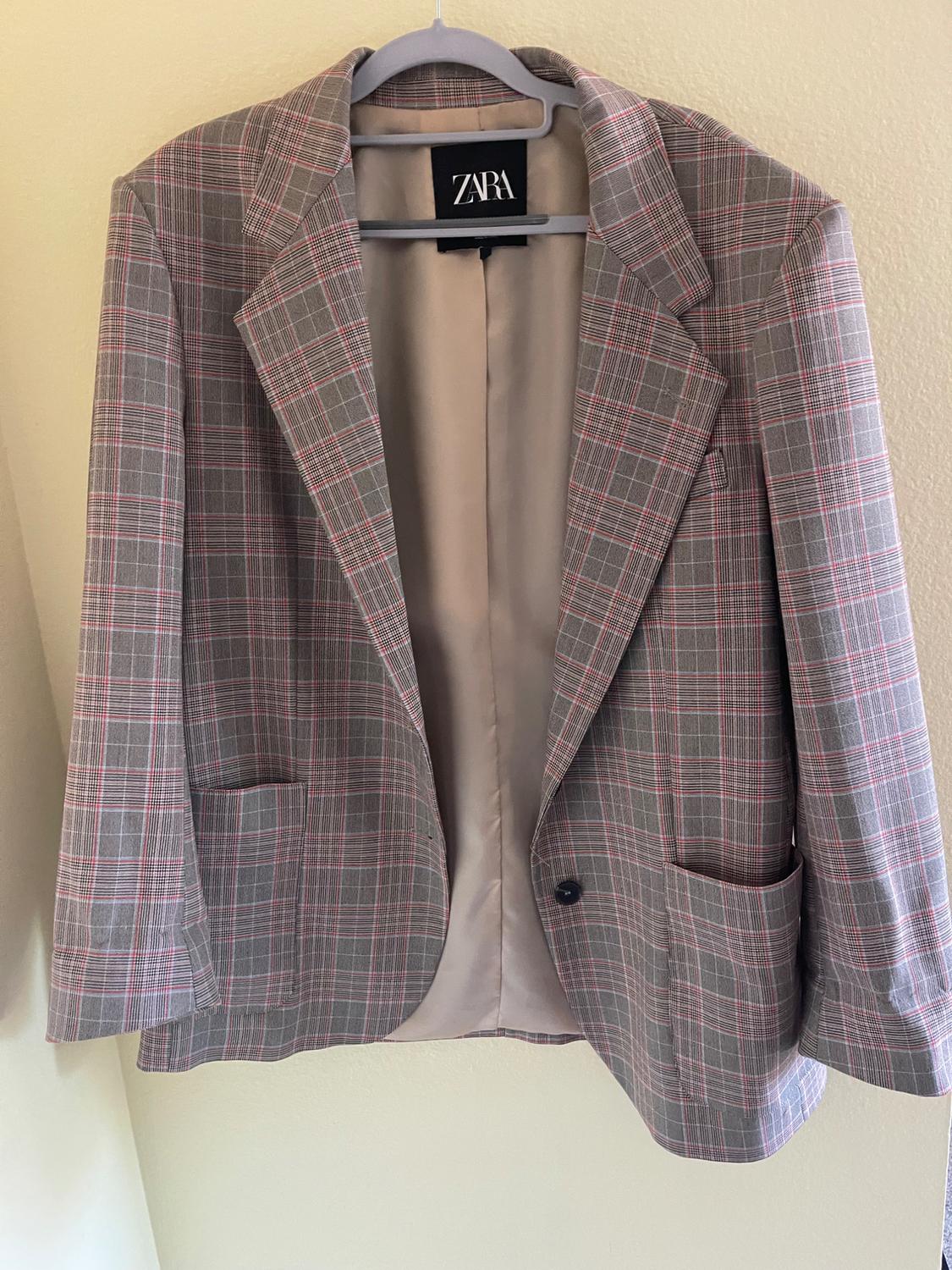 Grey and red checked jacket hanging on a hanger with a visible Zara tag, beige wall background.