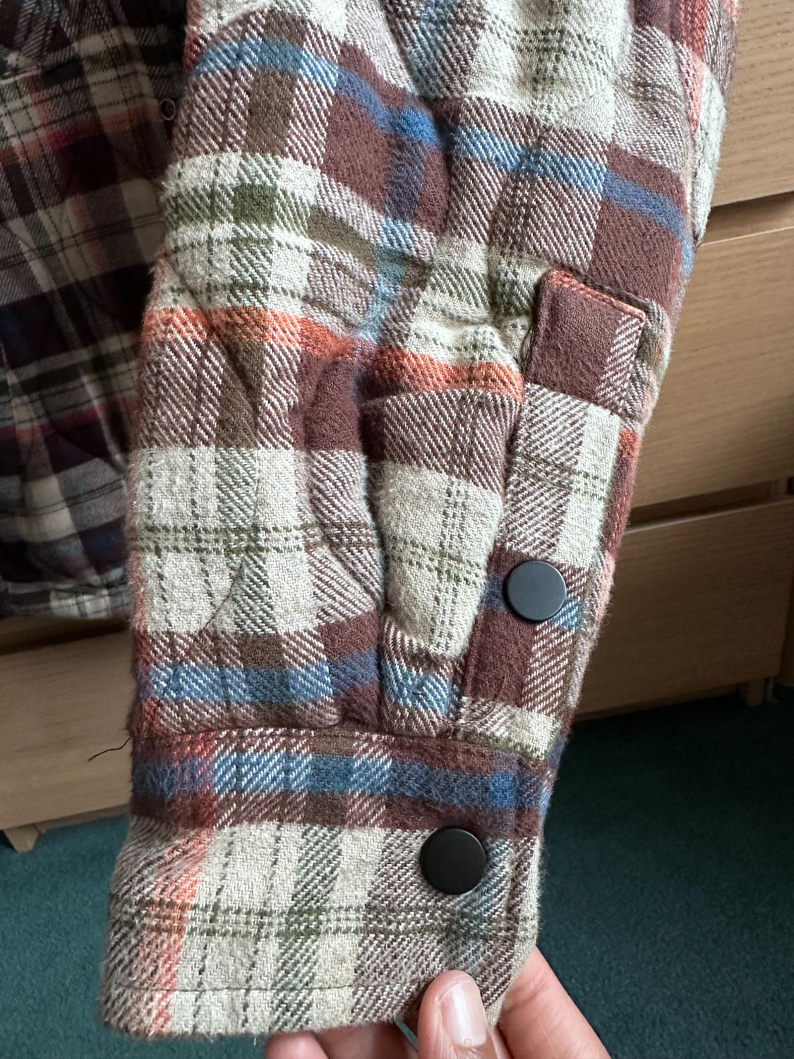 Checked shirt sleeve in brown, green, and blue tones with black buttons. Wooden background and green carpet. Hand holding the sleeve.