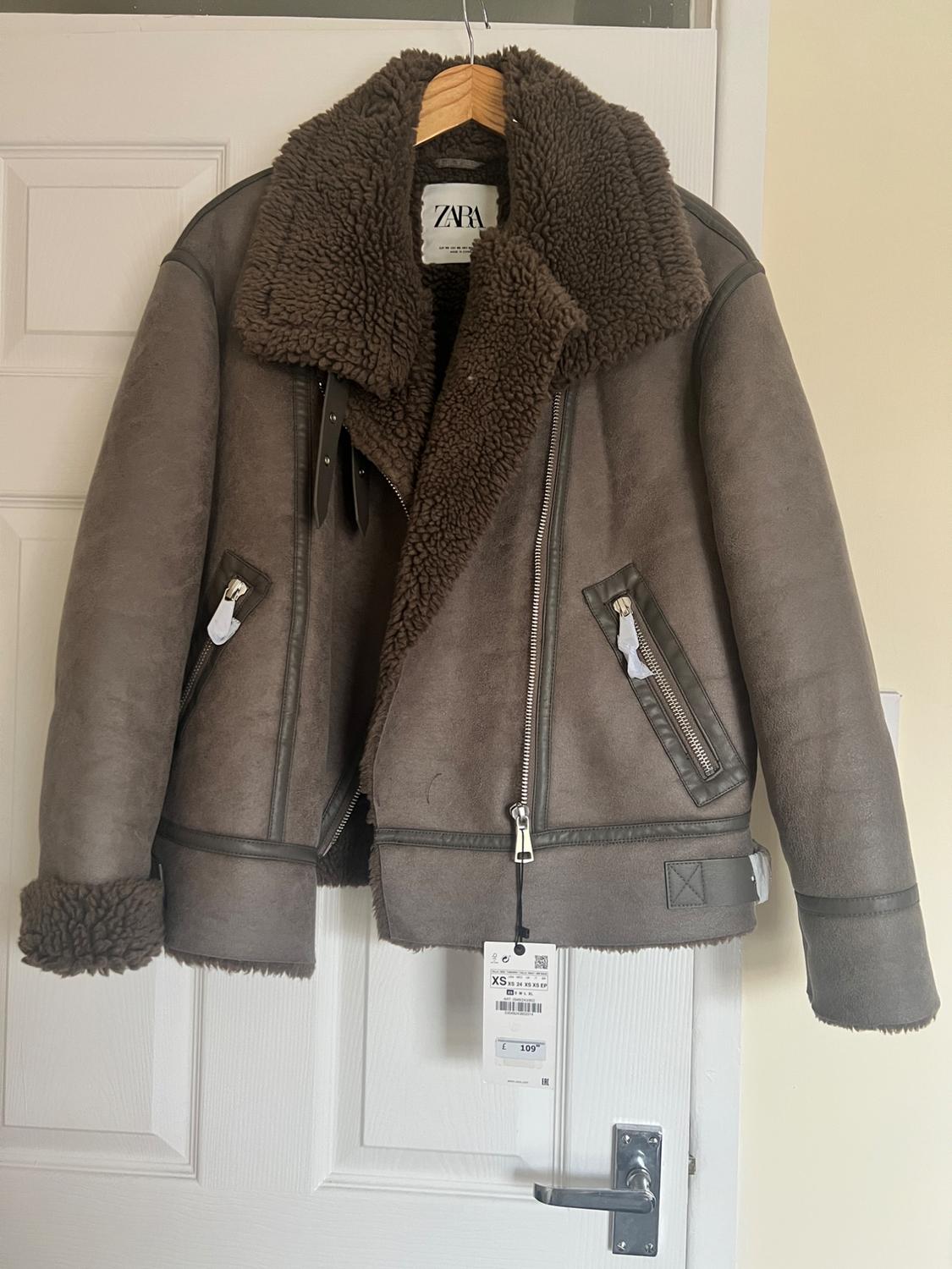 Brown jacket with sheepskin lining hanging on a white door. Visible tag with price of 109.95. ZARA brand.