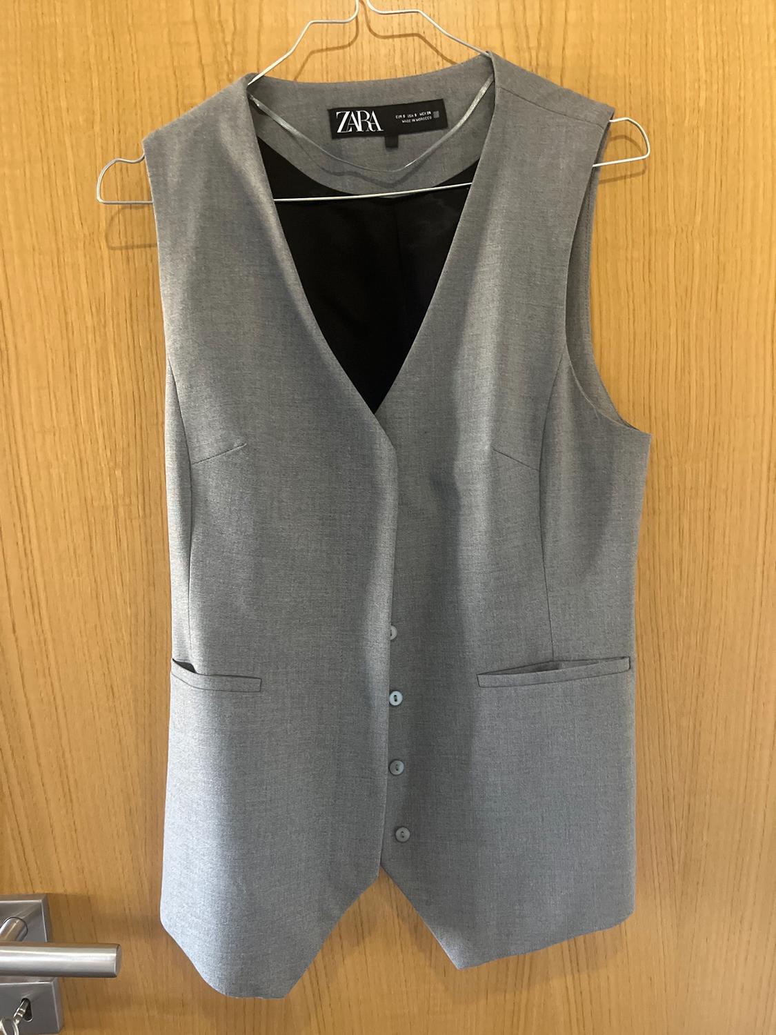 Grey dress waistcoat hanging on a hanger over a wooden door. Visible tag: ZARA.