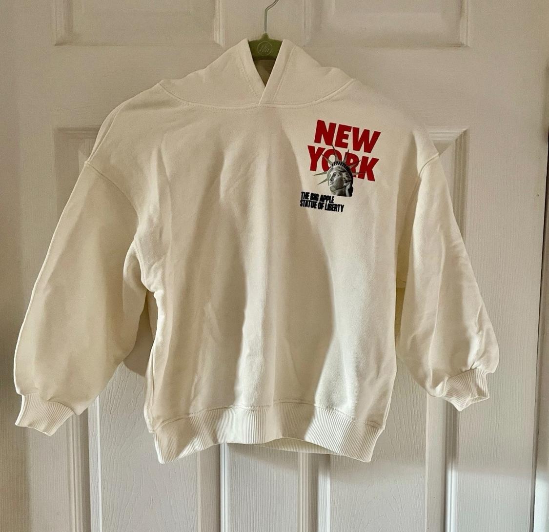 White sweatshirt hanging on a door, with "NEW YORK" text in red and "THE BIG APPLE STATUE OF LIBERTY" next to an image of the Statue of Liberty.