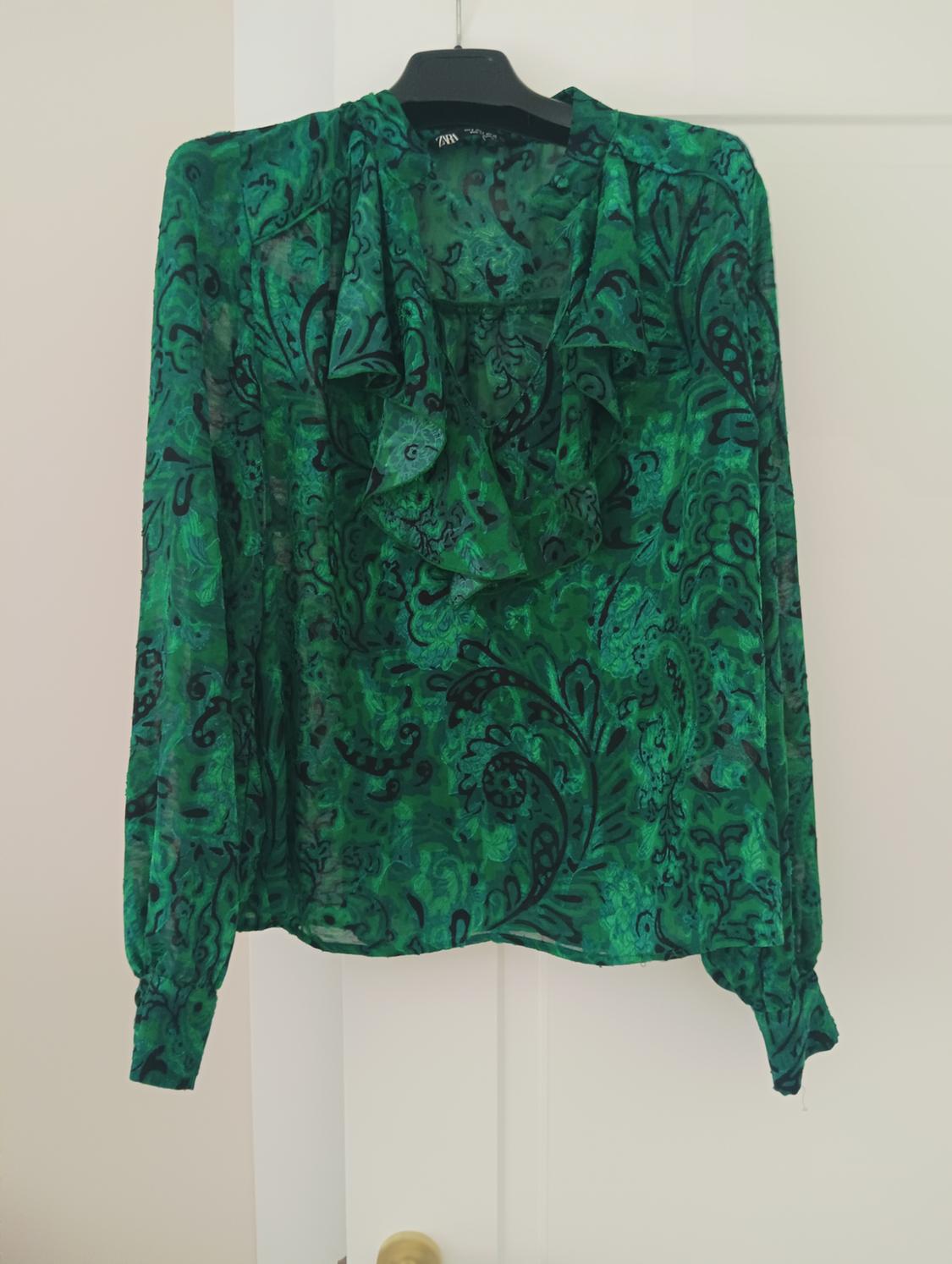 Green blouse with a black floral print, long sleeves, and a ruffled collar, hanging on a black hanger in front of a white door.