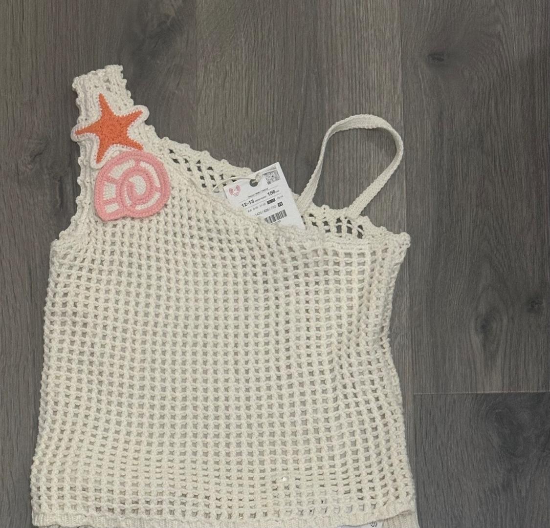 White crochet top with thin straps, decorated with orange starfish and pink snail patches, on a gray wooden background.