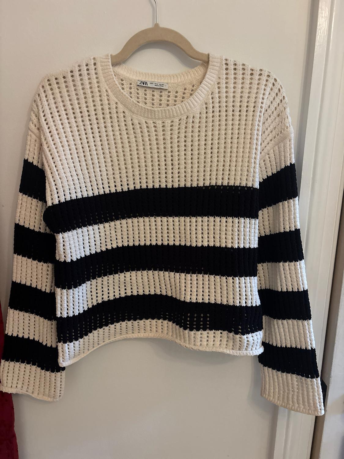 Black and white open-knit sweater hanging on a hanger on a white door. The label reads the text zara.