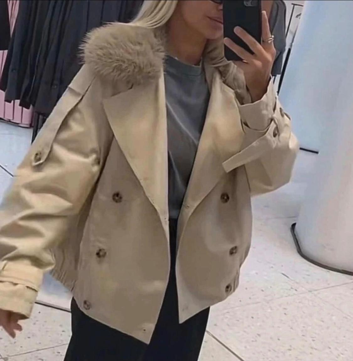 Woman taking a selfie in a shop mirror. She is wearing a beige double-breasted jacket with a fur collar, a grey T-shirt and black trousers.