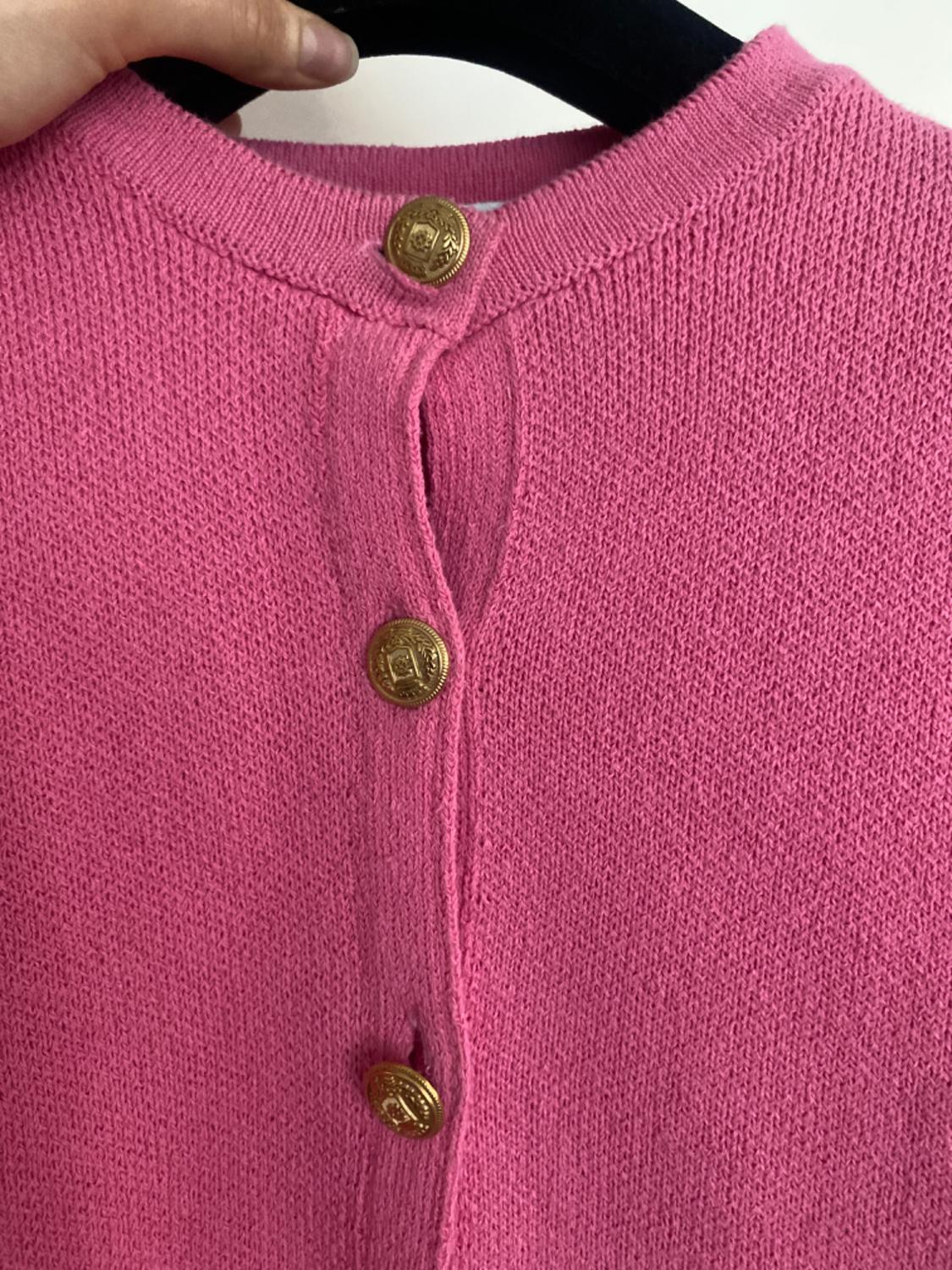Close-up of a pink knitted cardigan with a round neck and embossed gold buttons, hanging on a black hanger.