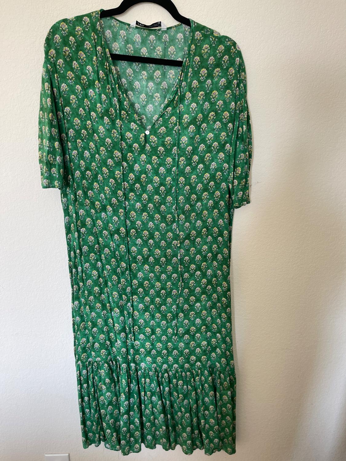 Green dress with white and yellow floral print, short sleeves and a V-neck, hanging on a black hanger against a white wall.