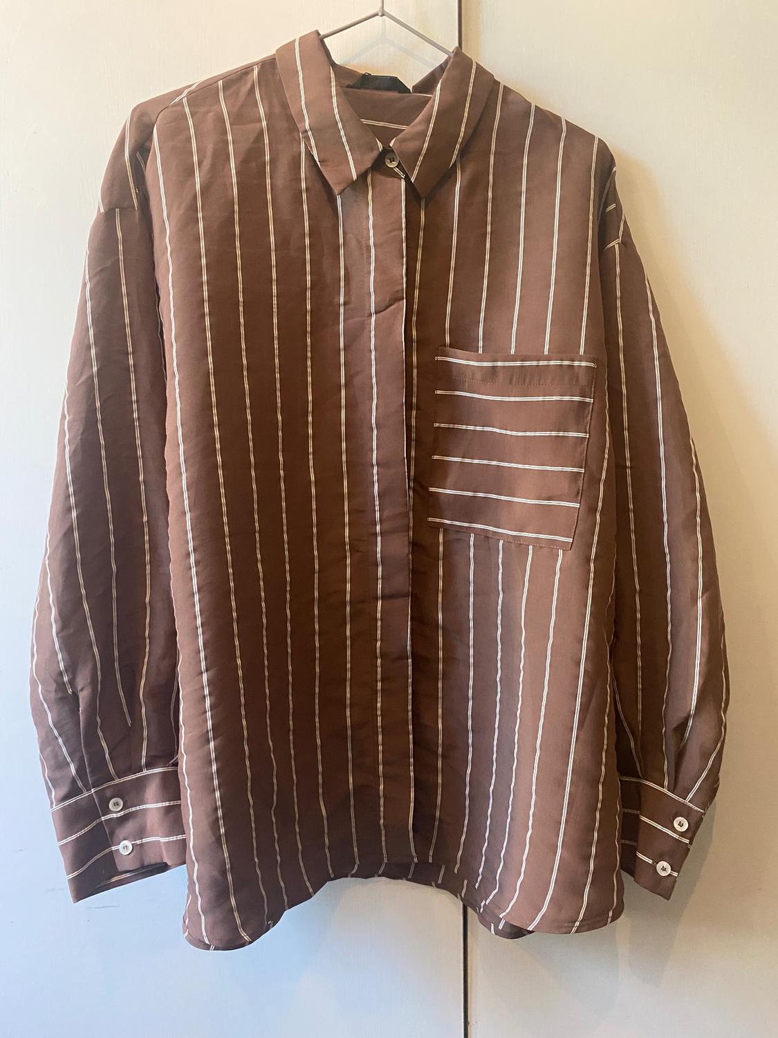 Brown shirt with white vertical stripes, with a chest pocket and buttons on the cuffs, hanging on a hanger.