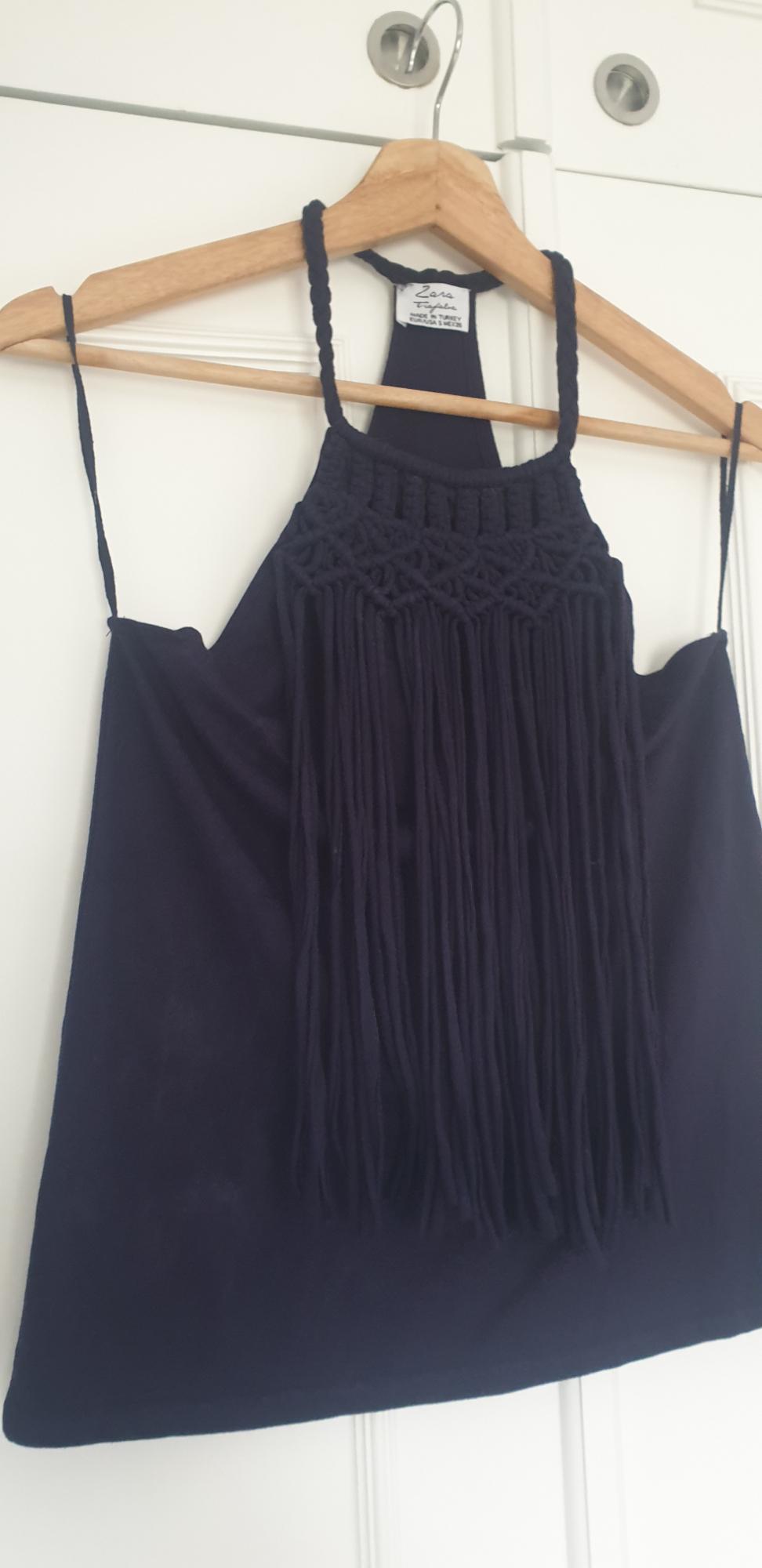 Black fringed top hanging on a wooden hanger. Visible label with text: "Zara Trafaluc". White background.