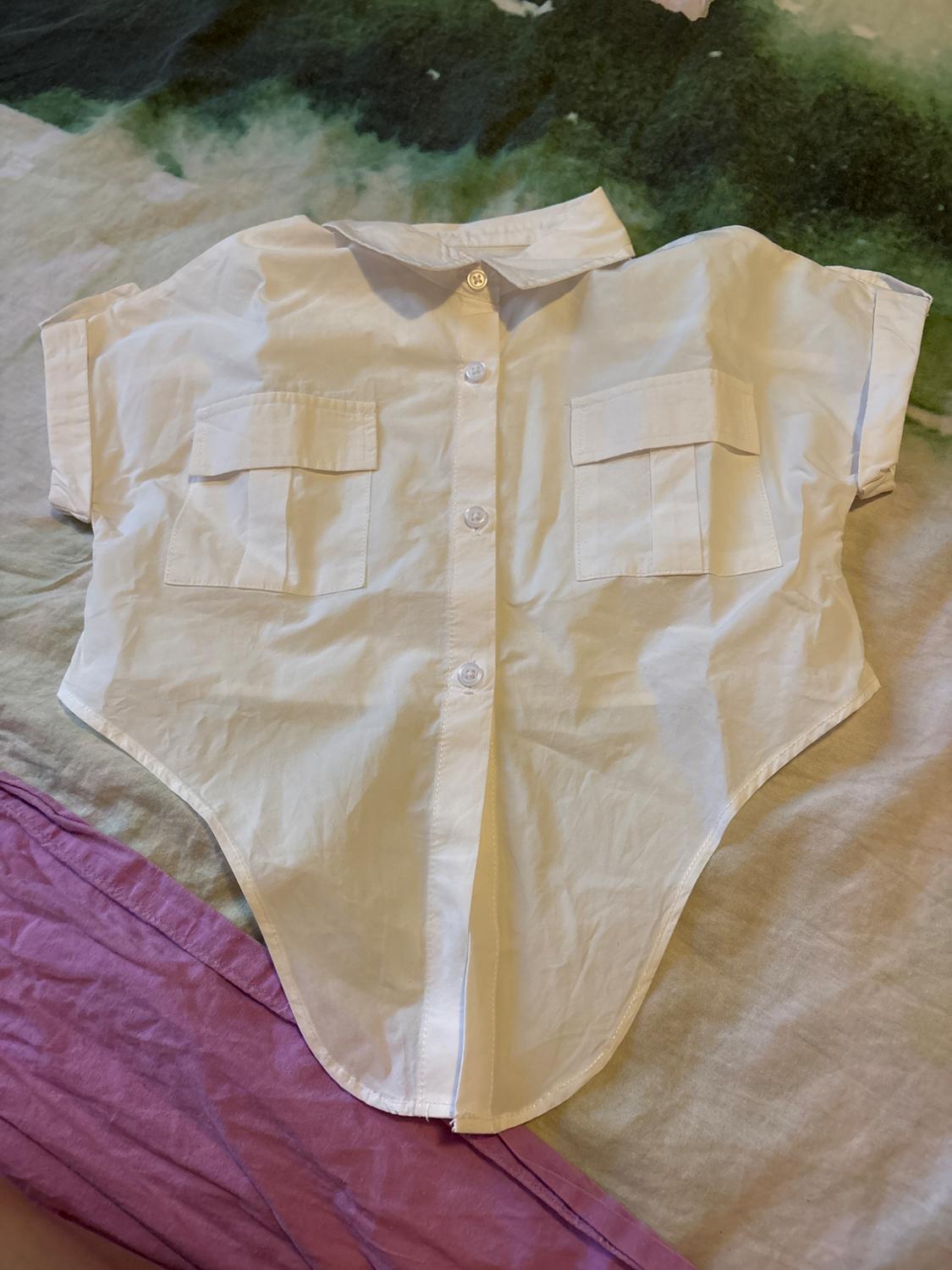 Short-sleeved white shirt with two front pockets, placed on a fabric with green and purple tones.