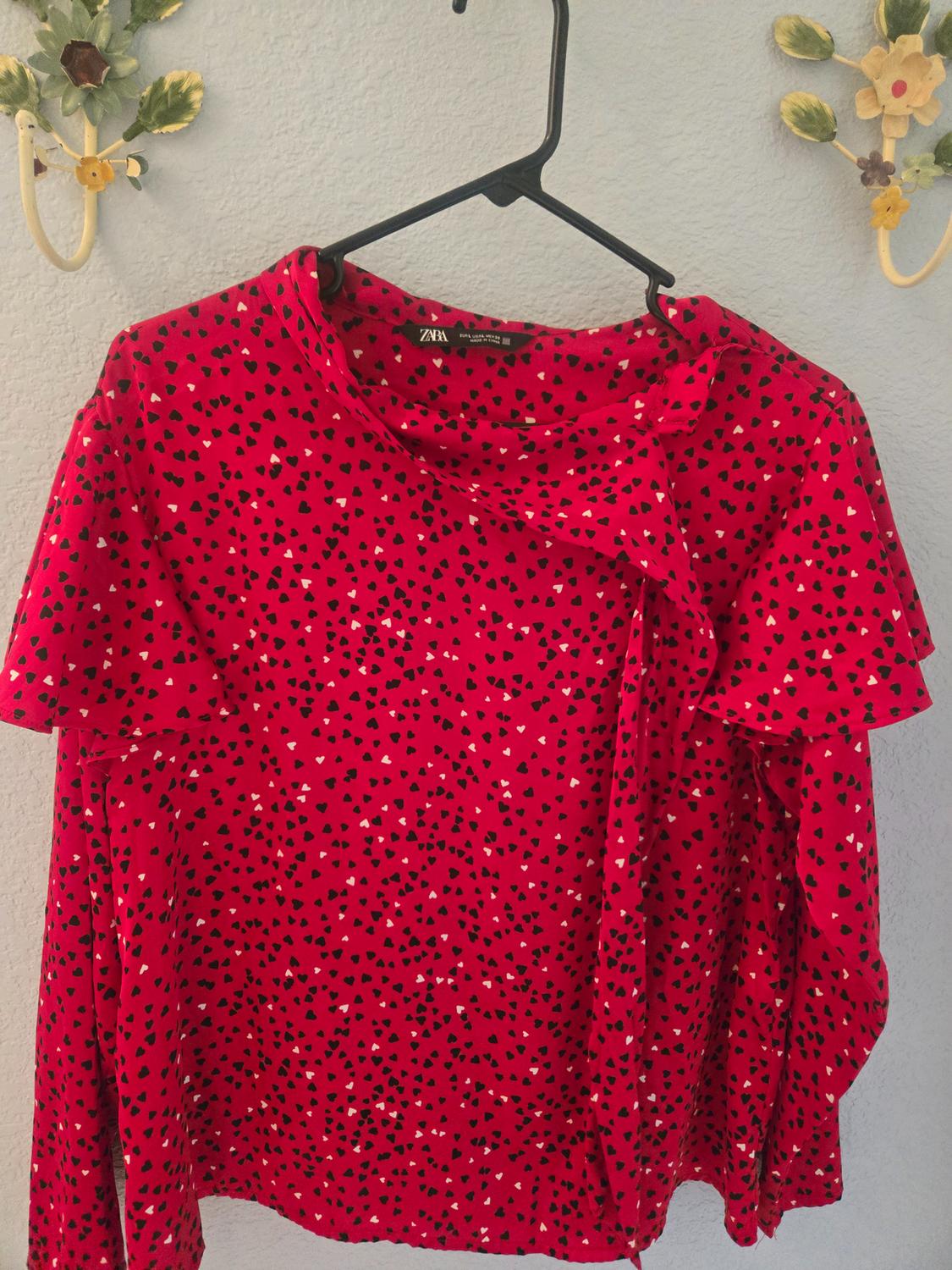 Red blouse with black and white heart print, hanging on a black hanger. Background with a light wall and floral decorations.