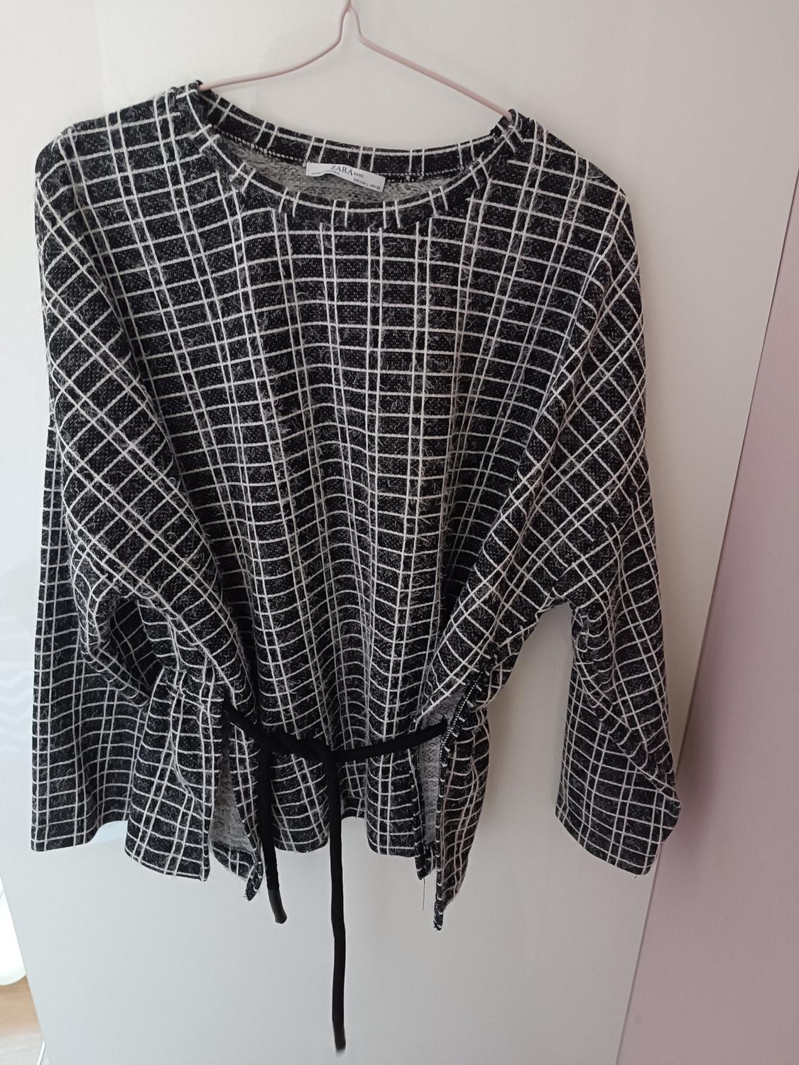 Black and white checked blouse hanging on a pink hanger. It has a black belt and a visible label on the neck.