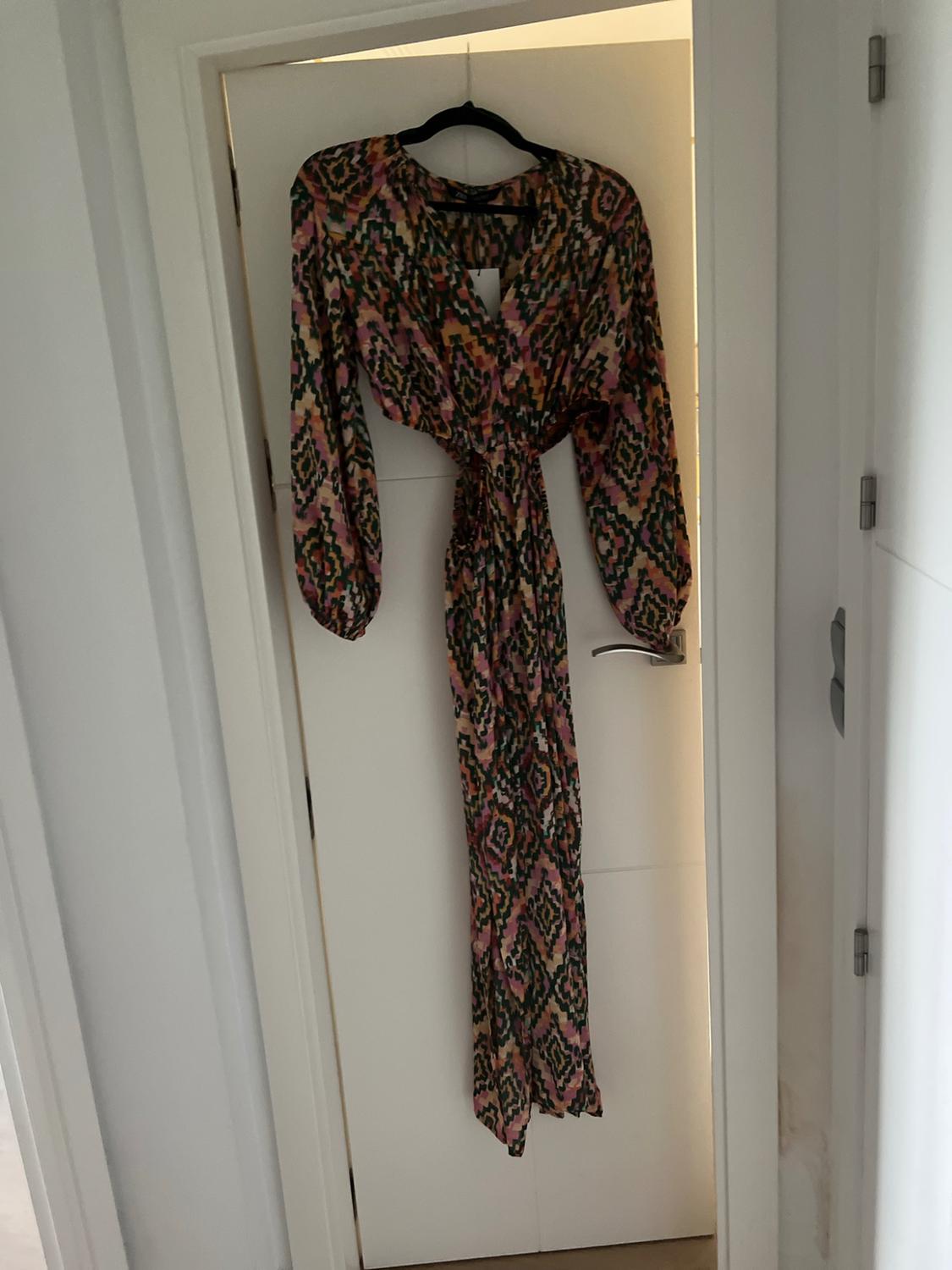 Maxi dress with a multicoloured geometric print, V-neck and cut-outs at the waist, hanging on a hanger on a white door.