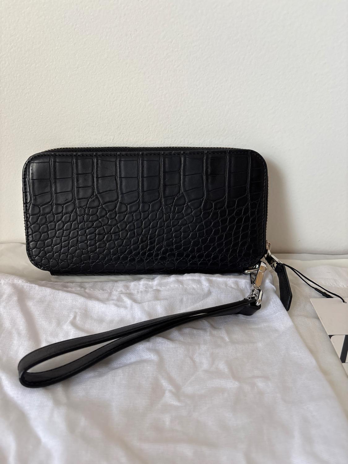 Black, leather-textured purse on white fabric, with a hand strap and metal clasp, resting on a light surface.