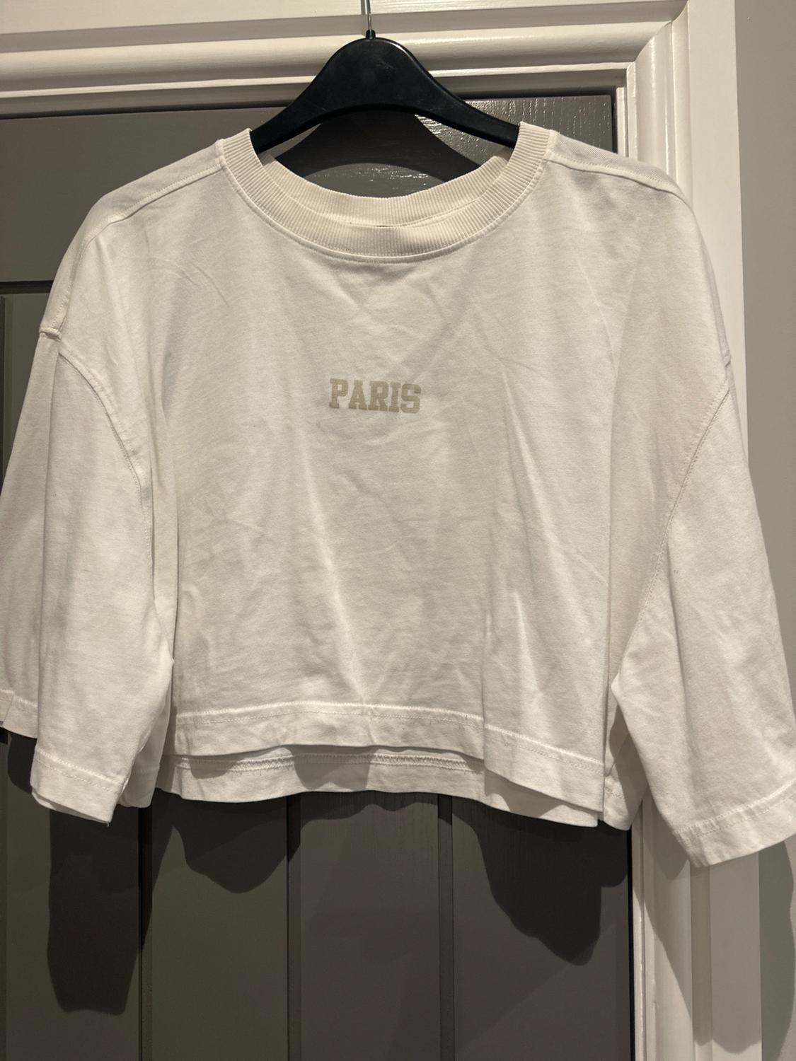 Short-sleeved white T-shirt hanging on a black coat hanger, with the word "PARIS" printed on the chest. Grey door background.