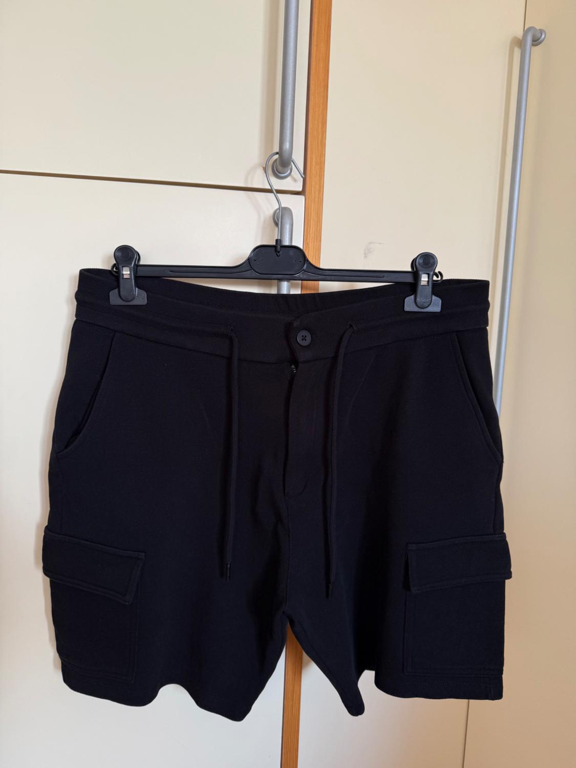Black shorts hanging on a hanger, with side pockets and an adjustable drawstring, against a beige wardrobe background.