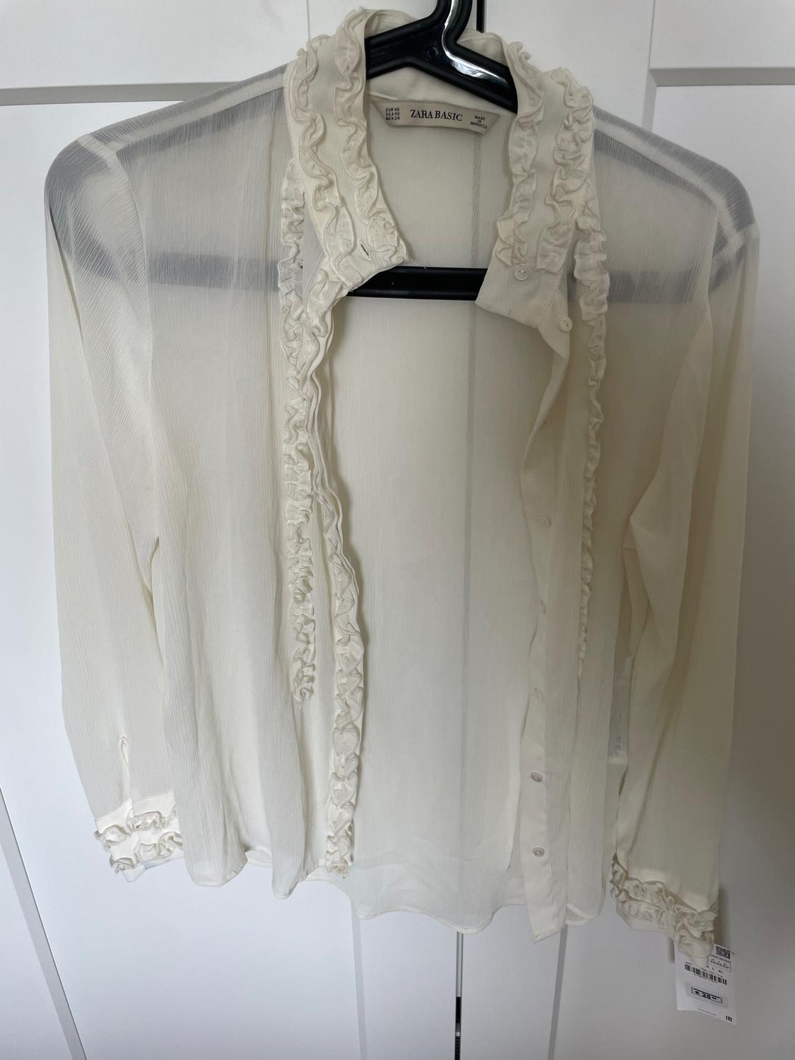 Semi-sheer white blouse with frills on the collar and cuffs, hanging on a black hanger. "ZARA BASIC" label visible.