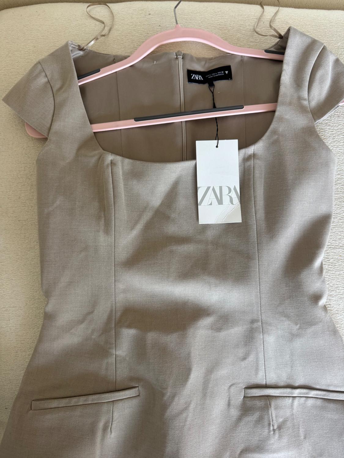 Beige short-sleeved dress on a pink hanger, with a Zara tag hanging. Light background and visible fabric texture.