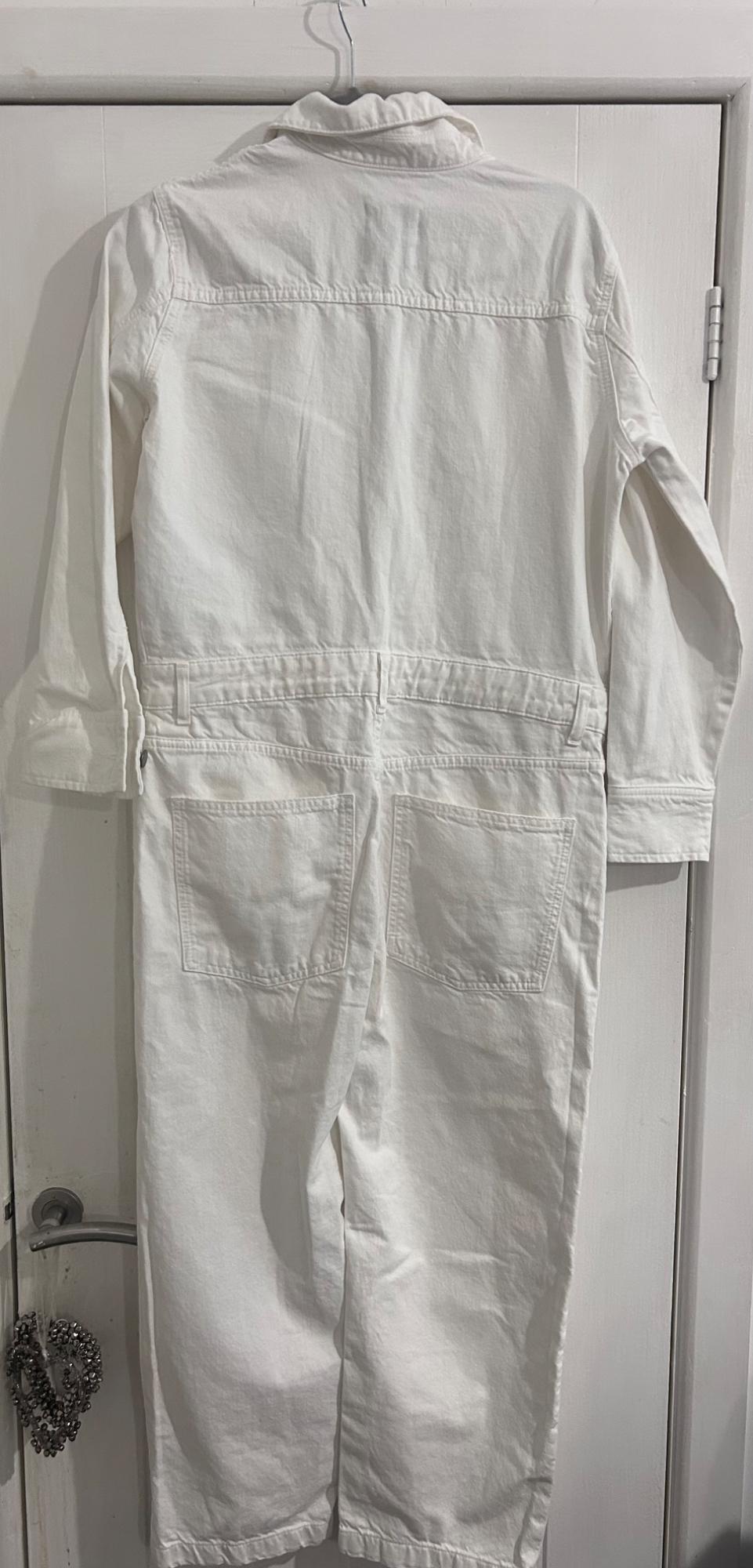 White jumpsuit hung on a white door. Seen from behind, with back pockets and long sleeves. Simple and minimalist setting.