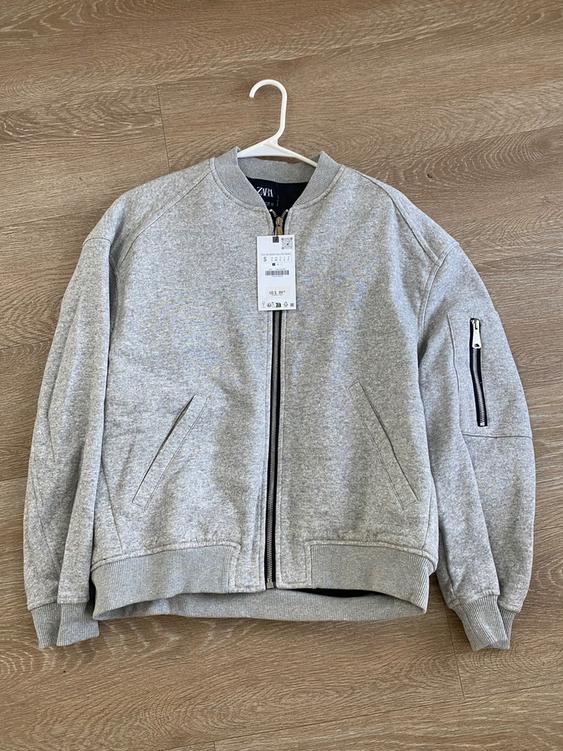 Gray jacket with zipper and sleeve pocket, hanging on a white hanger over a wooden floor. Visible tag.