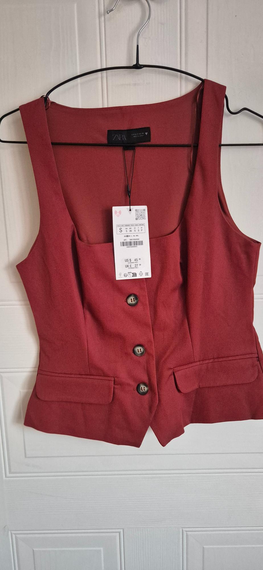 Dark red square-neck waistcoat with buttons hanging on a white door. Text: ZARA S US $ 45.90 UK £ 27.99.