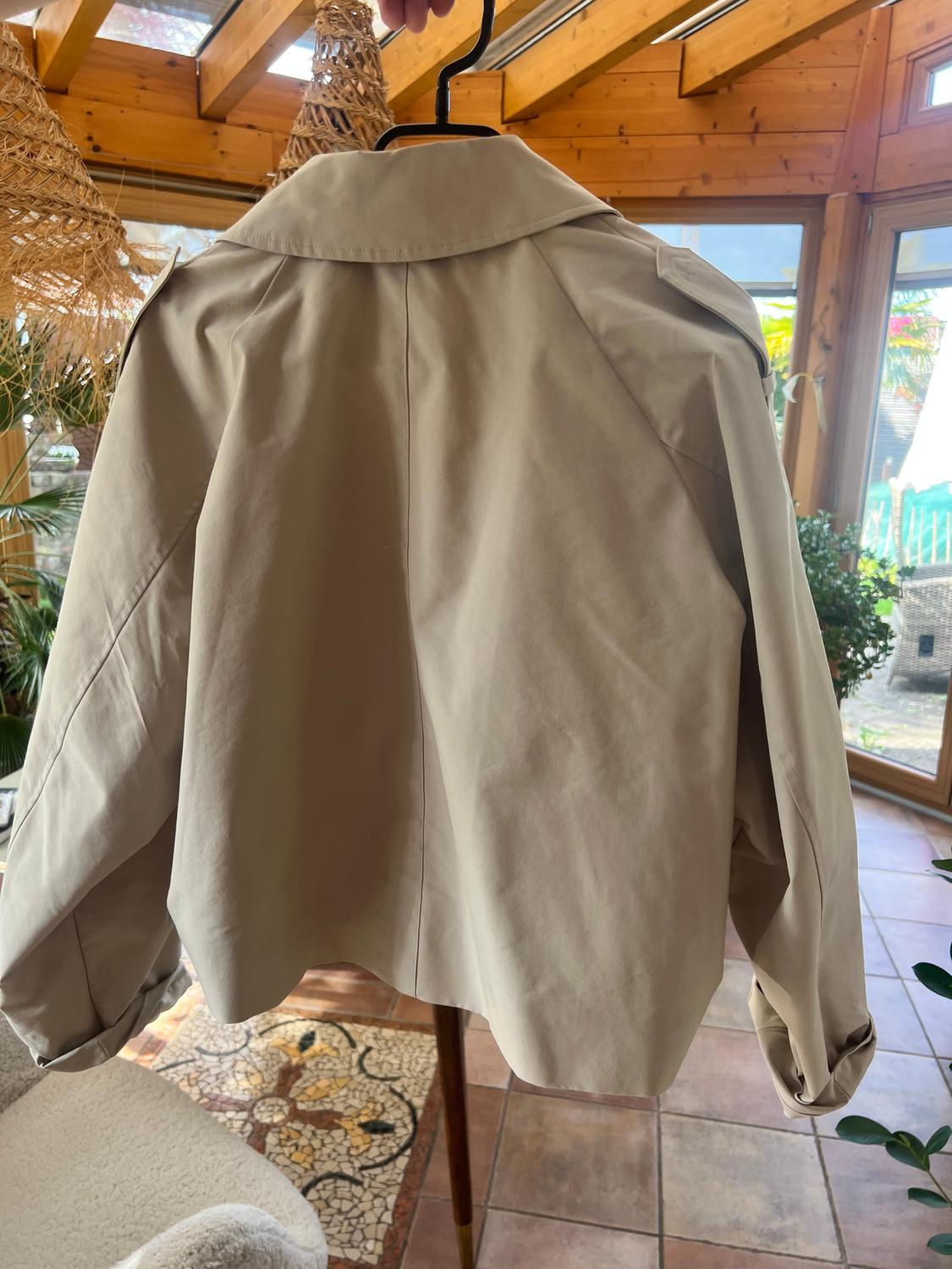 Beige jacket hanging on a hanger in a room with a wooden ceiling and large windows. Natural light illuminates the space.