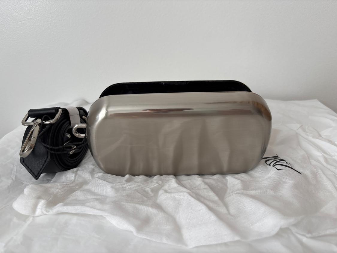 Rectangular metallic purse on white fabric, with a black strap coiled next to it. Plain white background.
